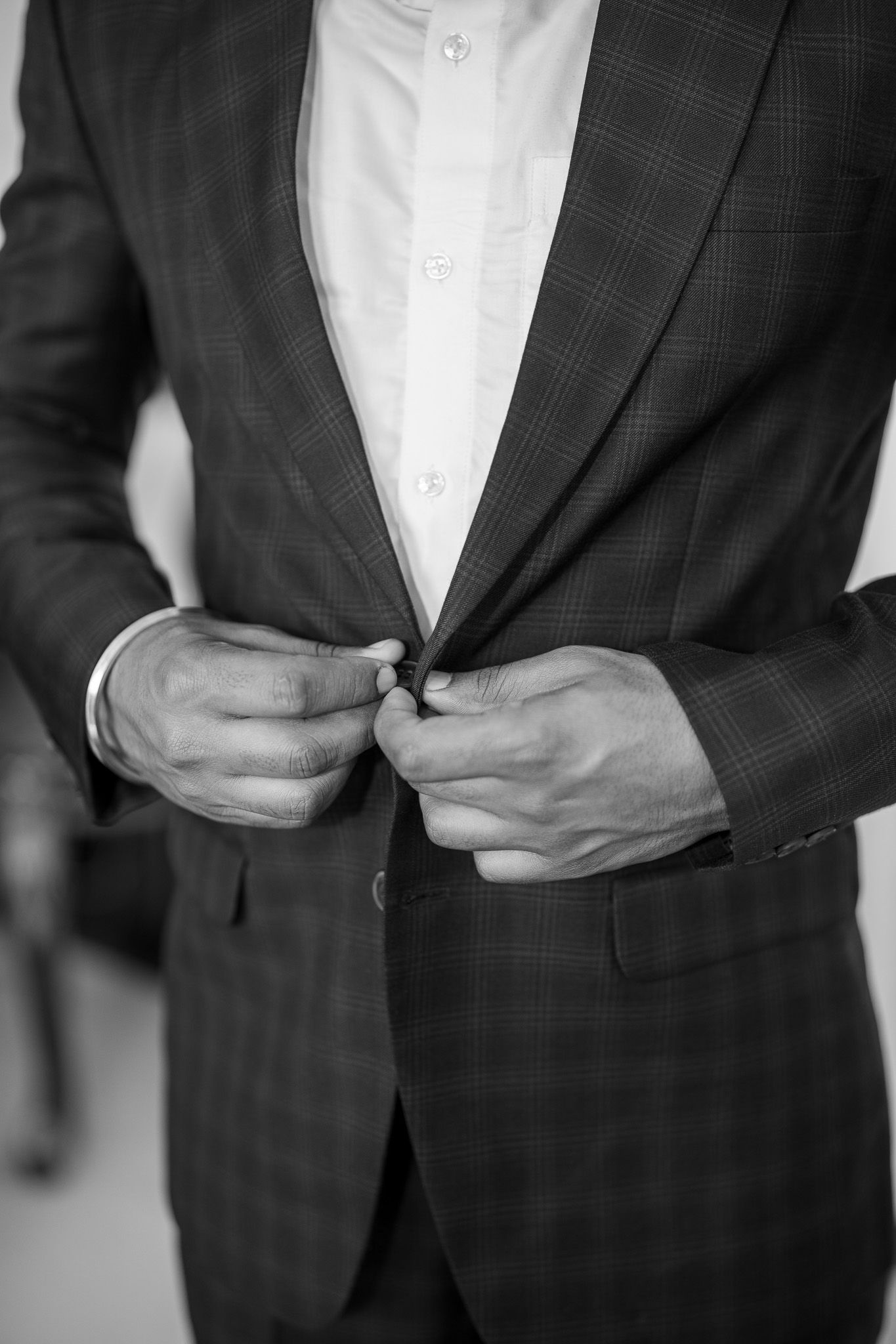 Close-up of man buttoning dark plaid suit jacket over white dress shirt, black and white photo.