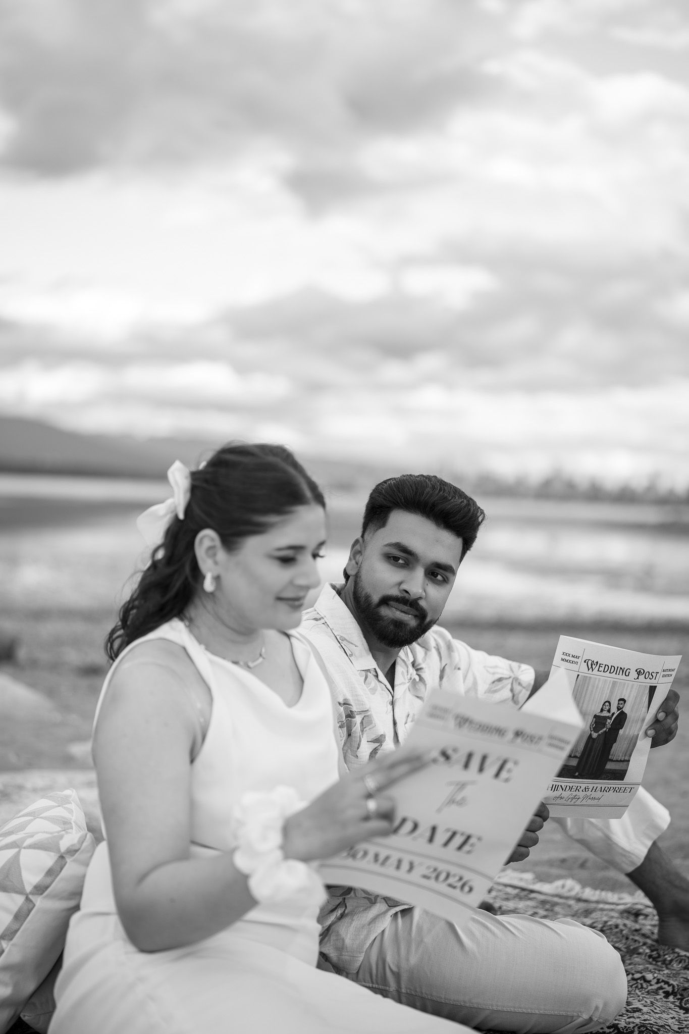 Black and white photo of couple reading Save the Date wedding newspaper on beach.