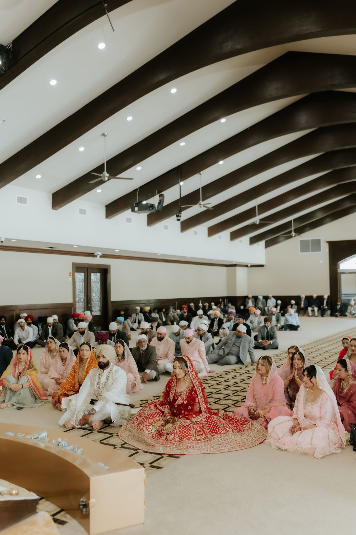 Sikh wedding ceremony in a gurdwara with bride in red lehenga seated among guests in colorful traditional attire.