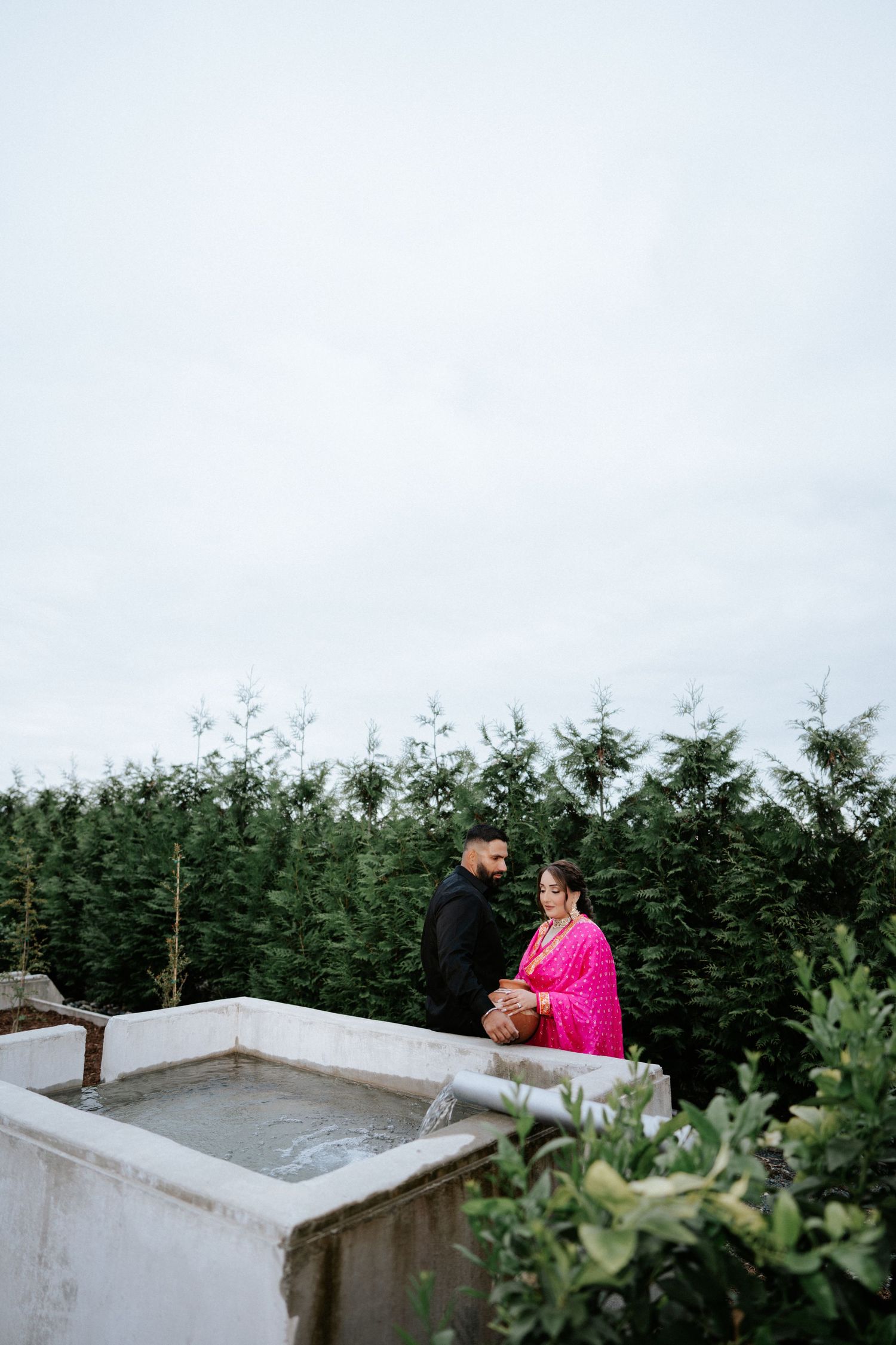 Couple in traditional South Asian attire standing on a rooftop surrounded by lush green trees on a cloudy day.