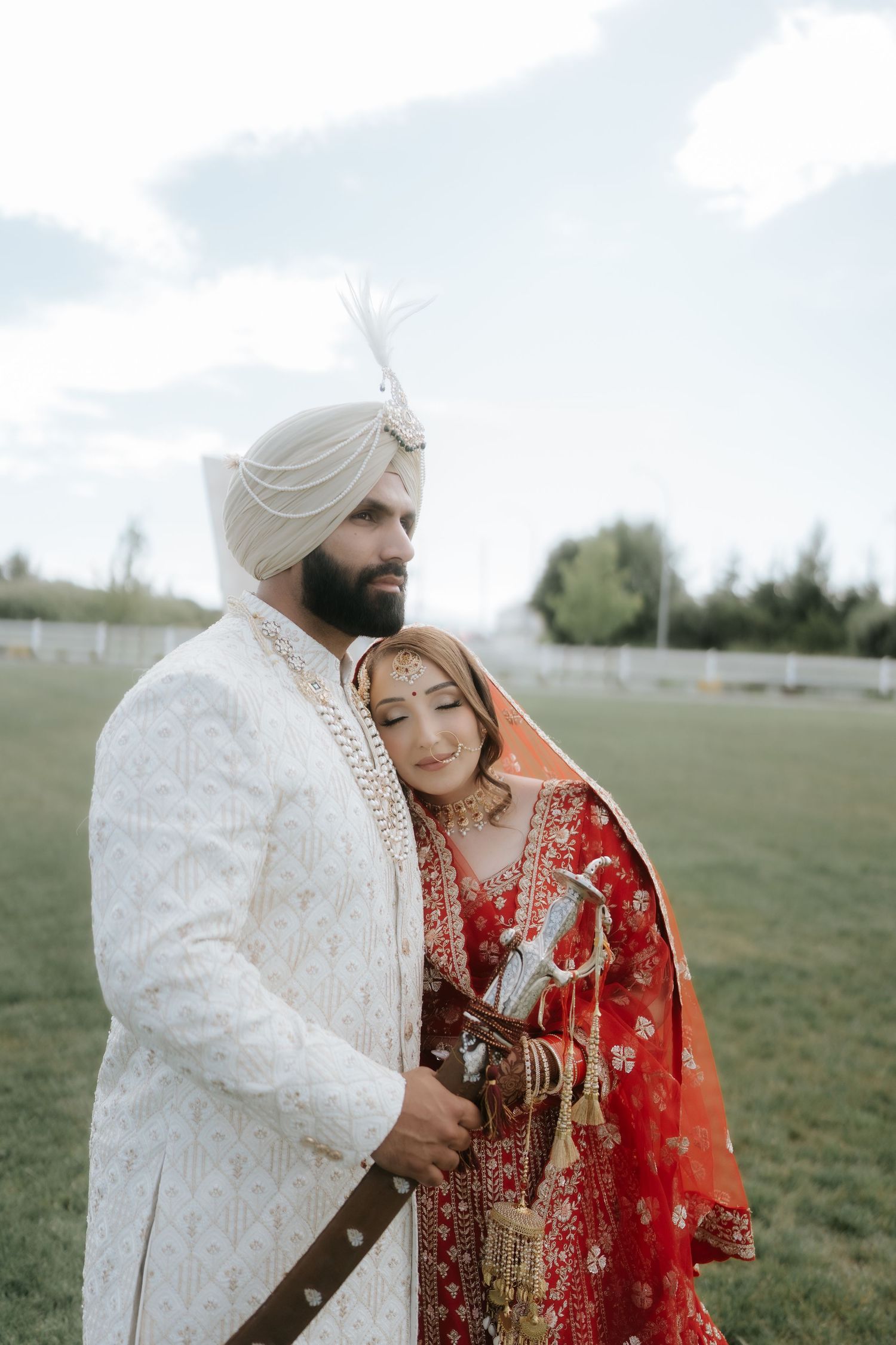 Sikh bride and groom in traditional wedding attire, groom in white sherwani and turban, bride in red lehenga outdoors.