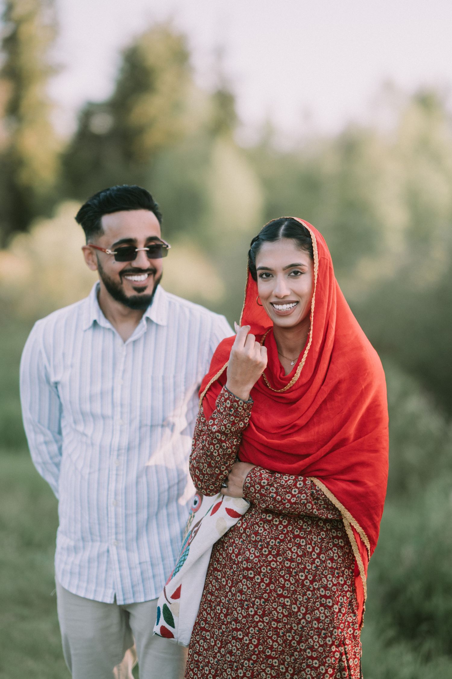 A couple shares joyful moments outdoors in traditional South Asian attire with the person wearing a bright red hijab.