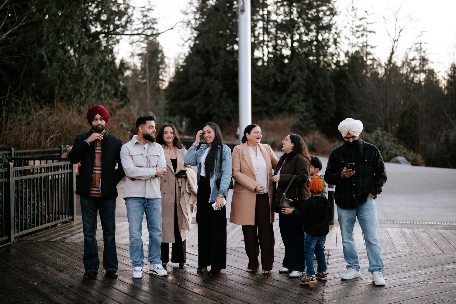 A group of people bundled up in winter clothing stand together on a wooden deck surrounded by evergreen trees.