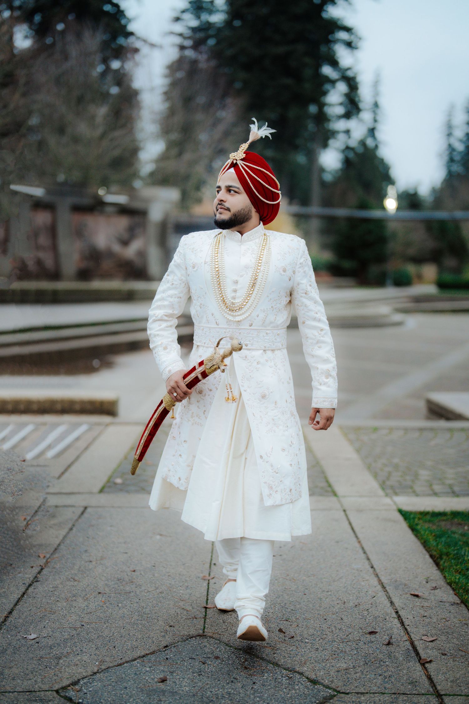 A person in white formal attire and red turban walks along outdoor concrete path.