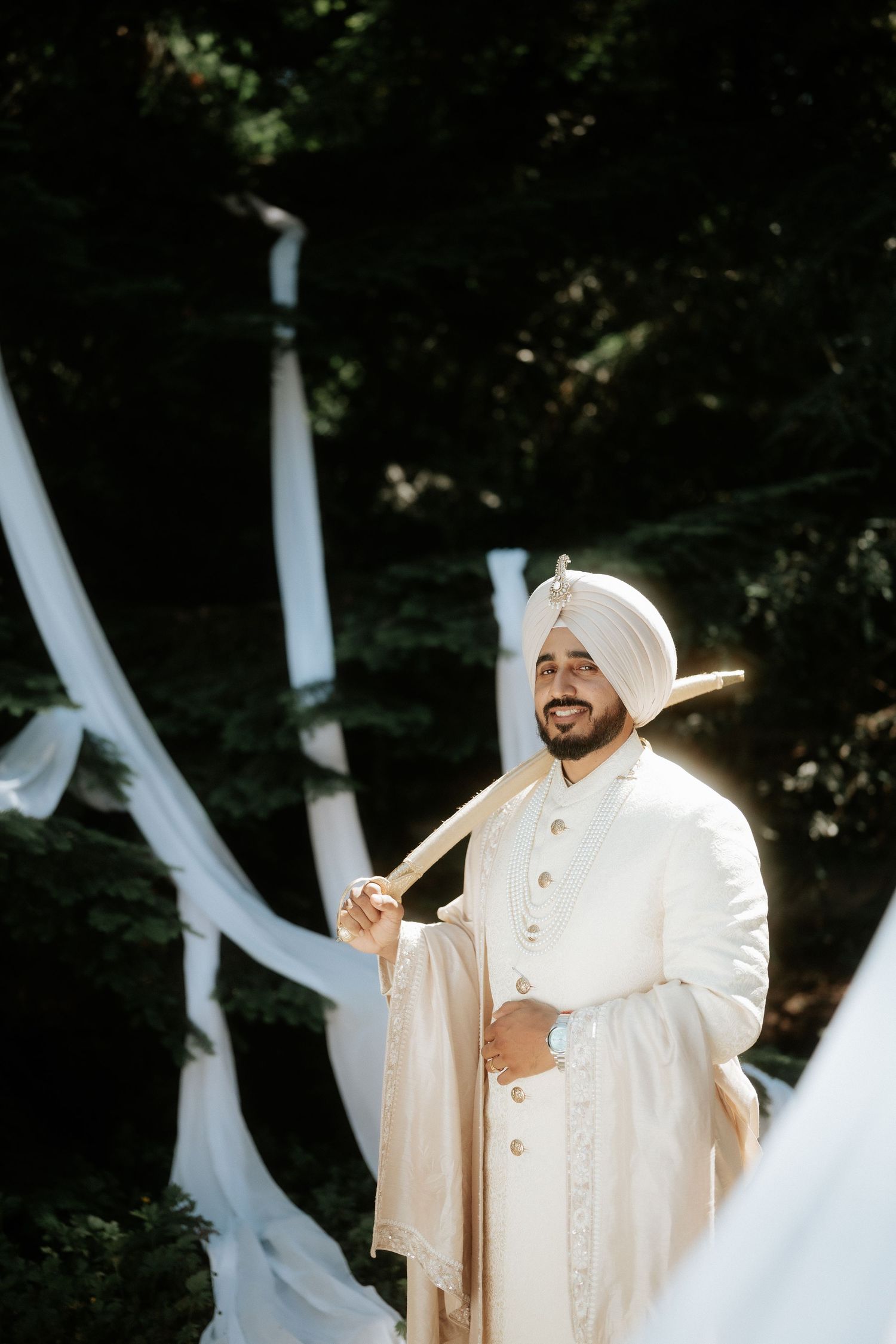 A groom in traditional white Sikh wedding attire poses against a backdrop of flowing white fabric and greenery.