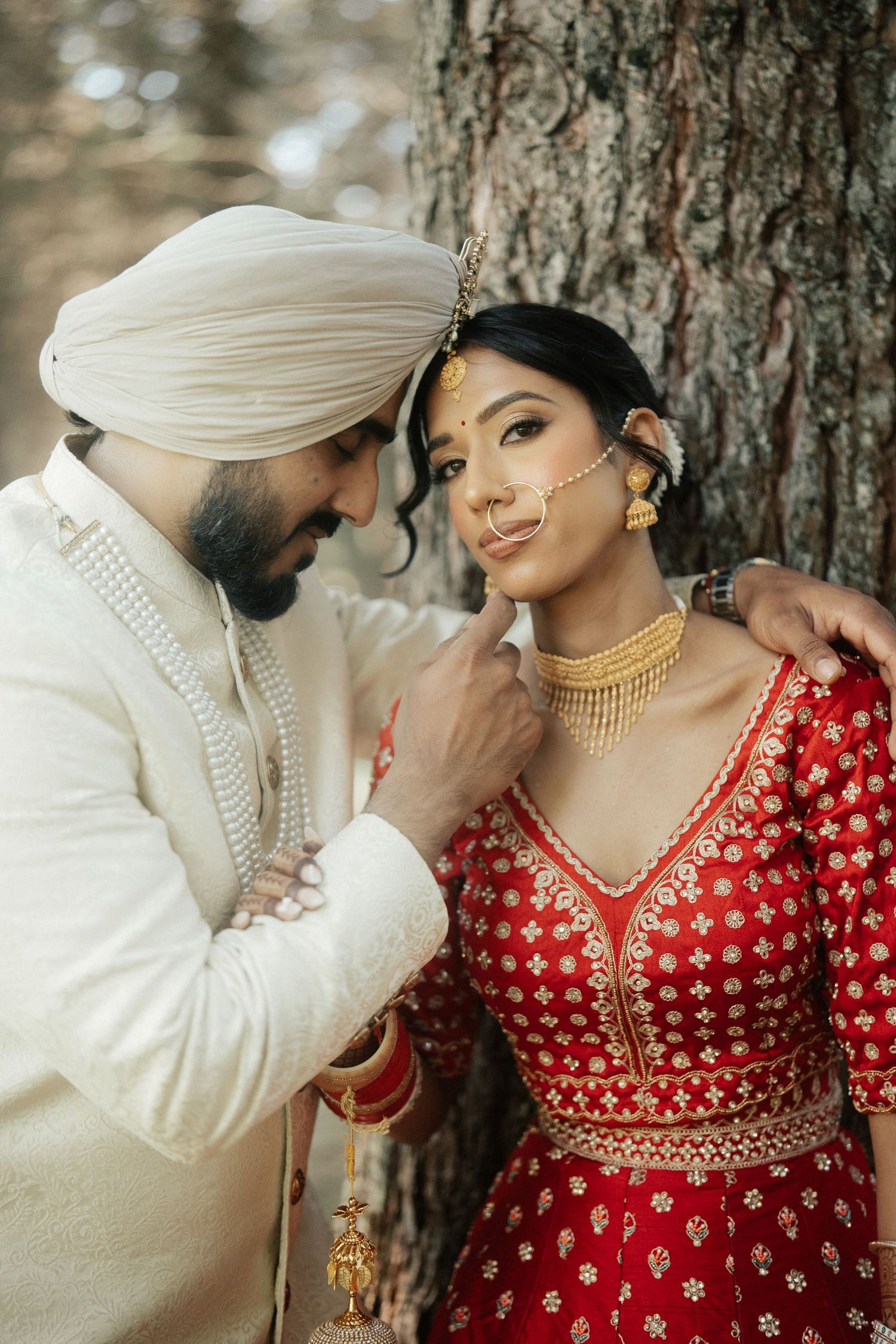 A romantic couple in traditional Indian wedding attire pose intimately against a tree backdrop.