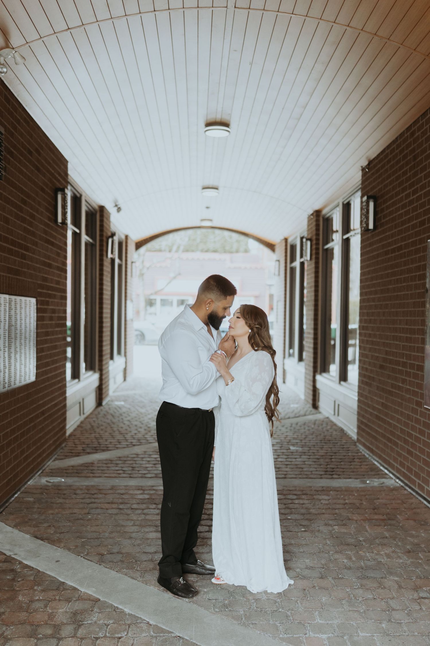 A couple in wedding attire embraces in a brick-lined covered walkway with white ceiling.
