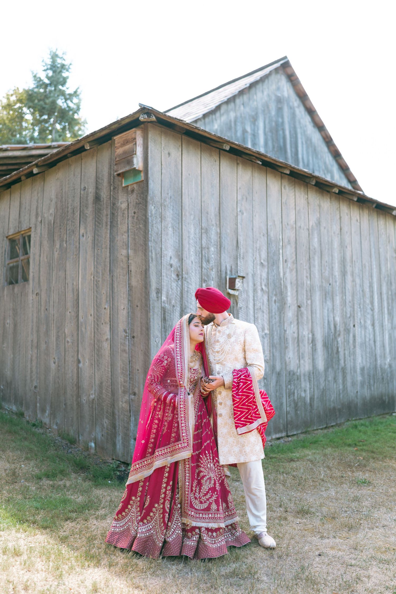 Sikh bride and groom in traditional red lehenga and cream sherwani sharing a kiss outside a rustic barn.