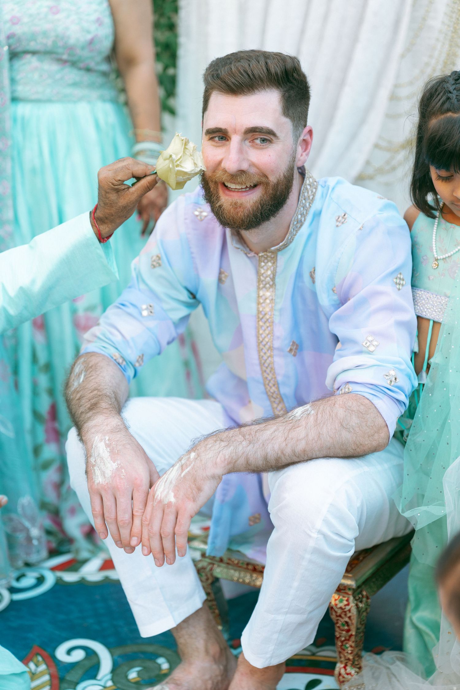 Smiling bearded man in light blue kurta and white pants during a traditional South Asian haldi ceremony.
