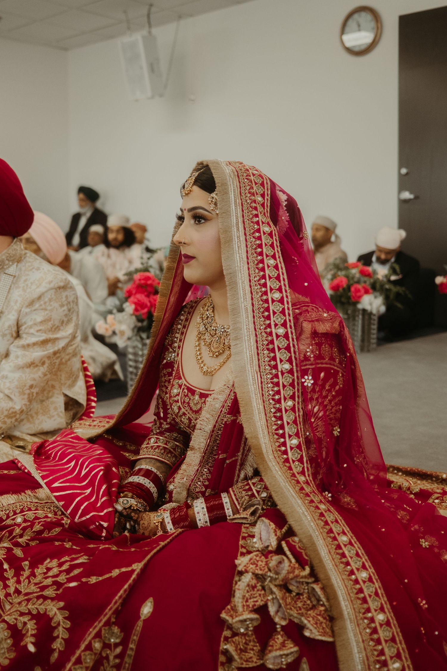 A Sikh bride in a red and gold lehenga with dupatta and traditional jewelry sits gracefully during her wedding ceremony.