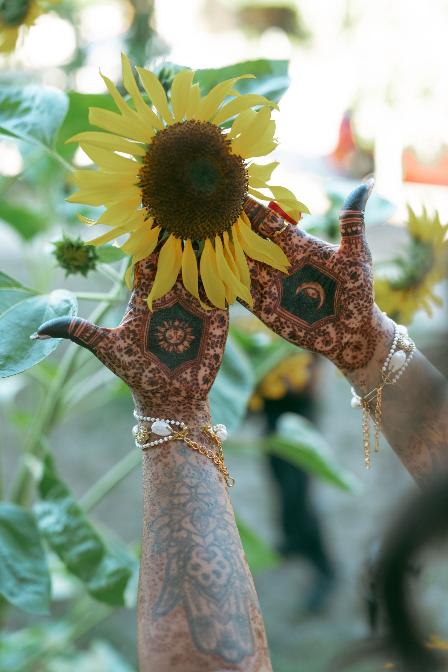 Hands adorned with intricate henna mehndi designs and bracelets framing a sunflower in a garden.