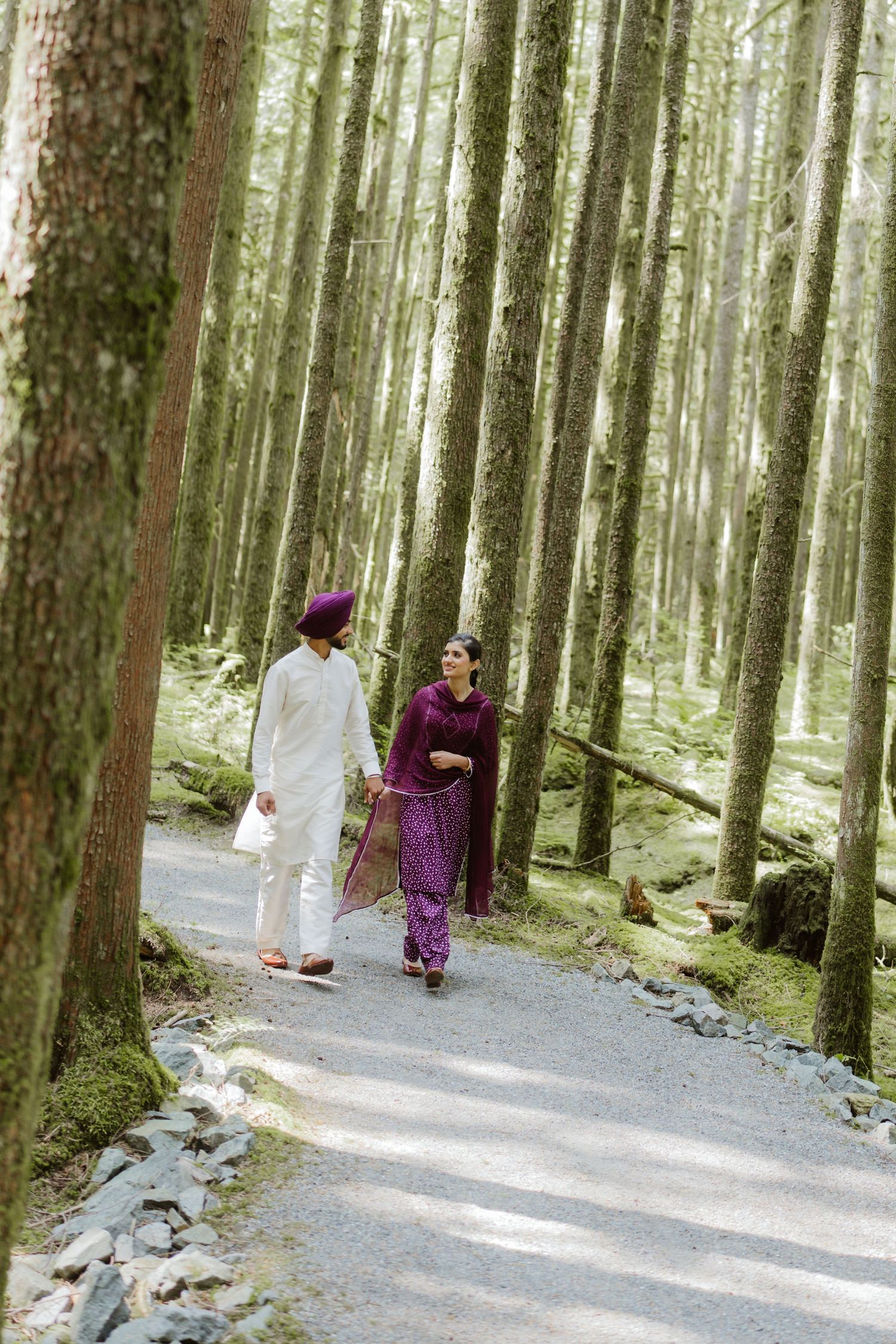 A Sikh couple in traditional purple and white attire walks hand in hand along a forest path.