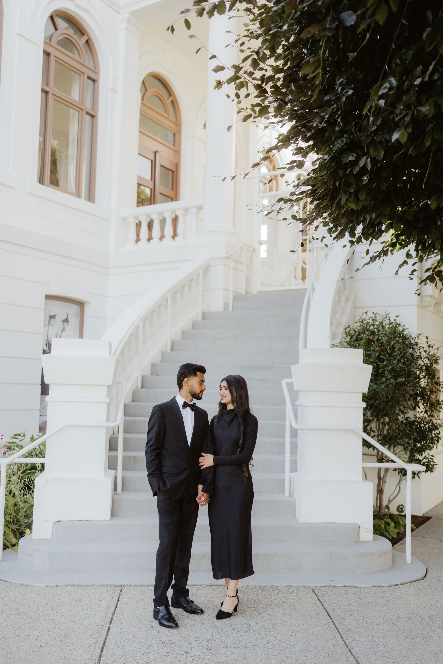 Elegant couple in matching black formal outfits holding hands in front of a white building staircase.