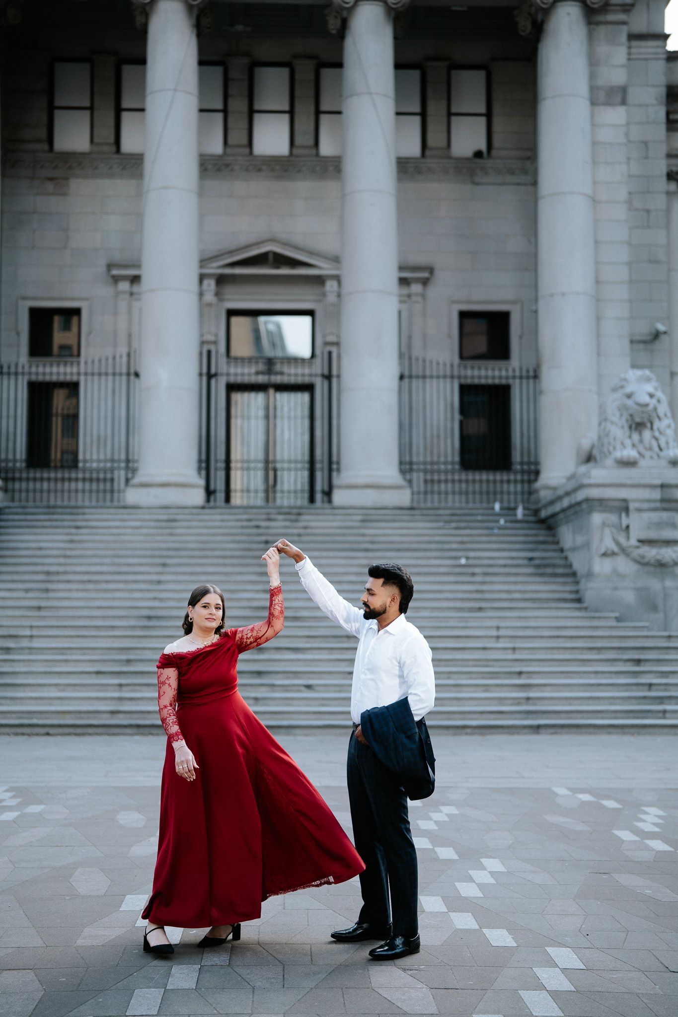 Couple dancing elegantly outside neoclassical building, woman in flowing red gown, man in white shirt and black trousers.