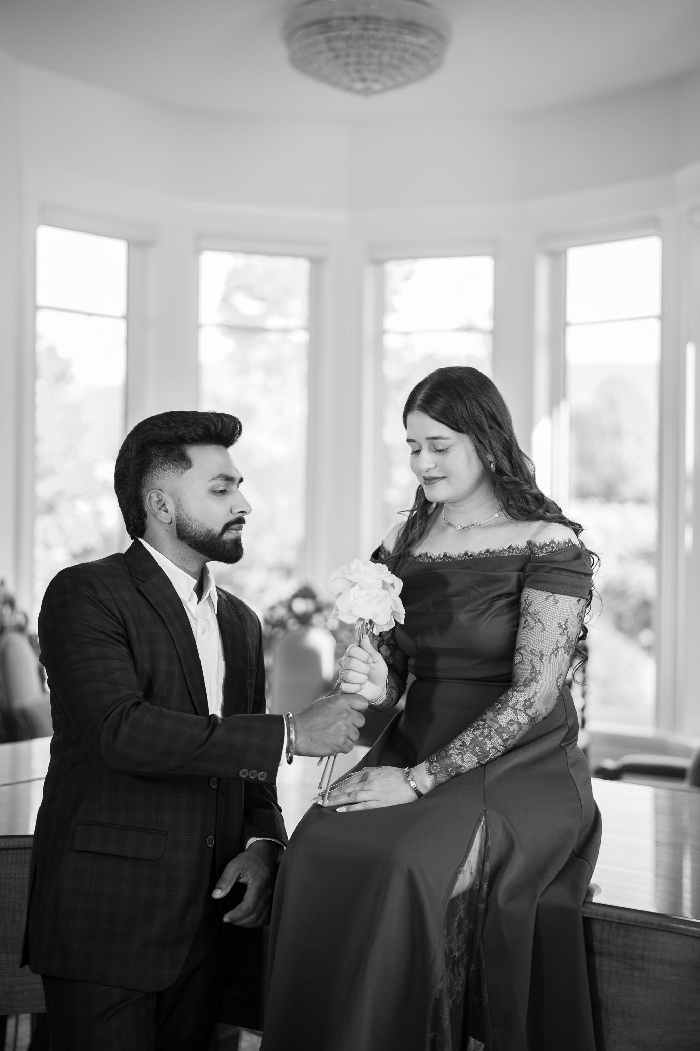 Black and white photo of a man in suit presenting white roses to a woman in an elegant off-shoulder gown.