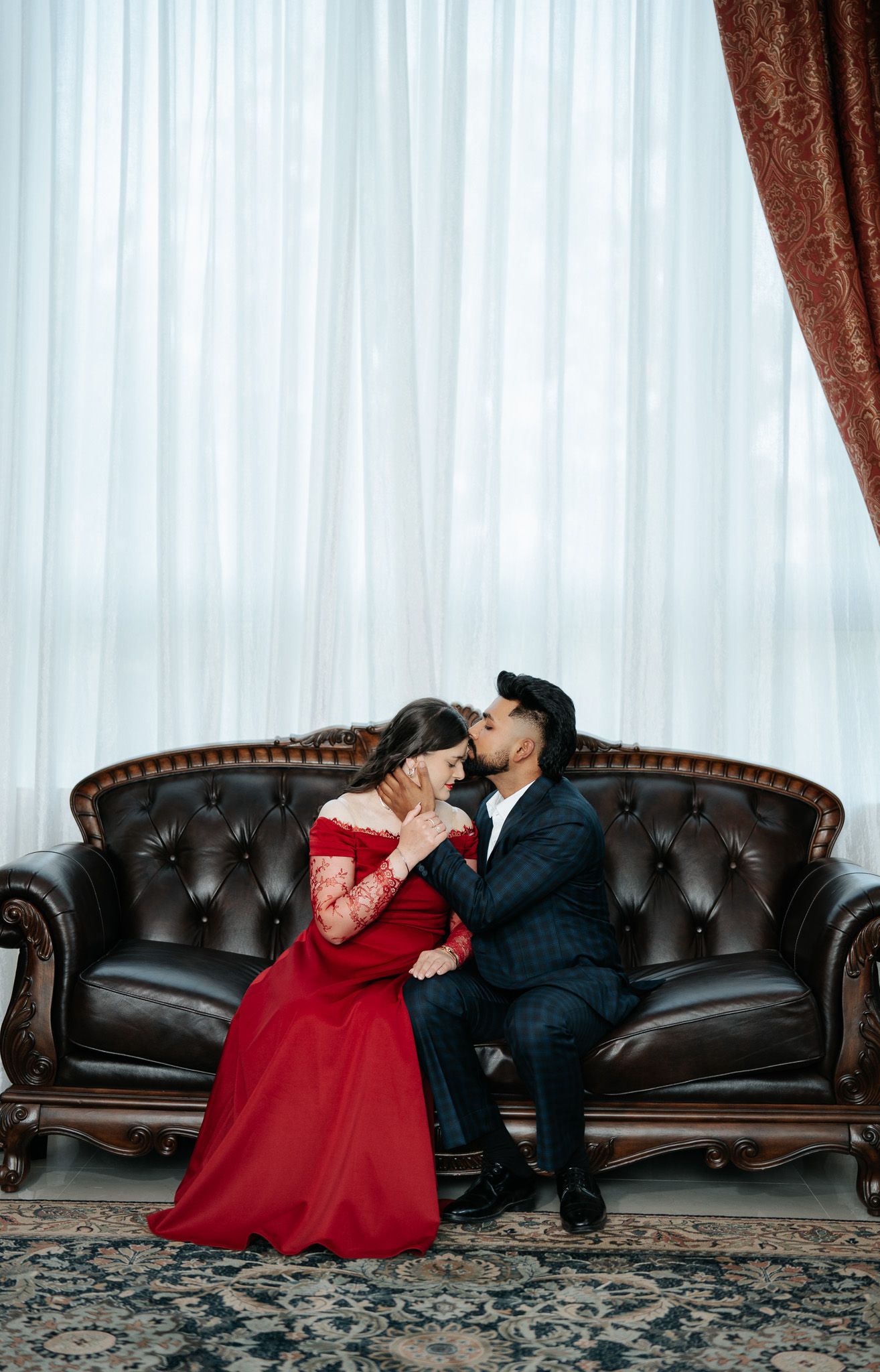 Couple sharing a romantic kiss on a tufted leather sofa, woman in red gown, man in dark suit, sheer curtains behind.