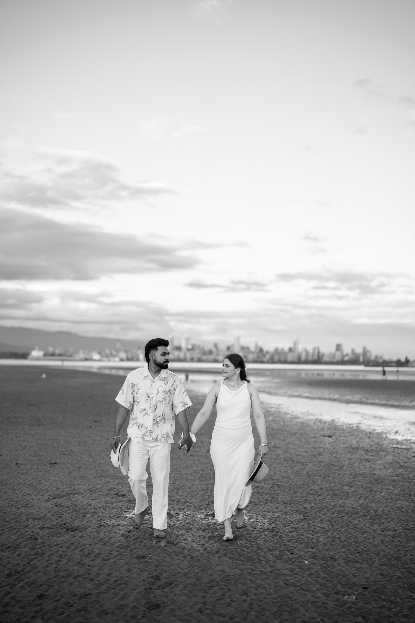 A couple walks hand in hand barefoot on a beach with a city skyline visible in the background.
