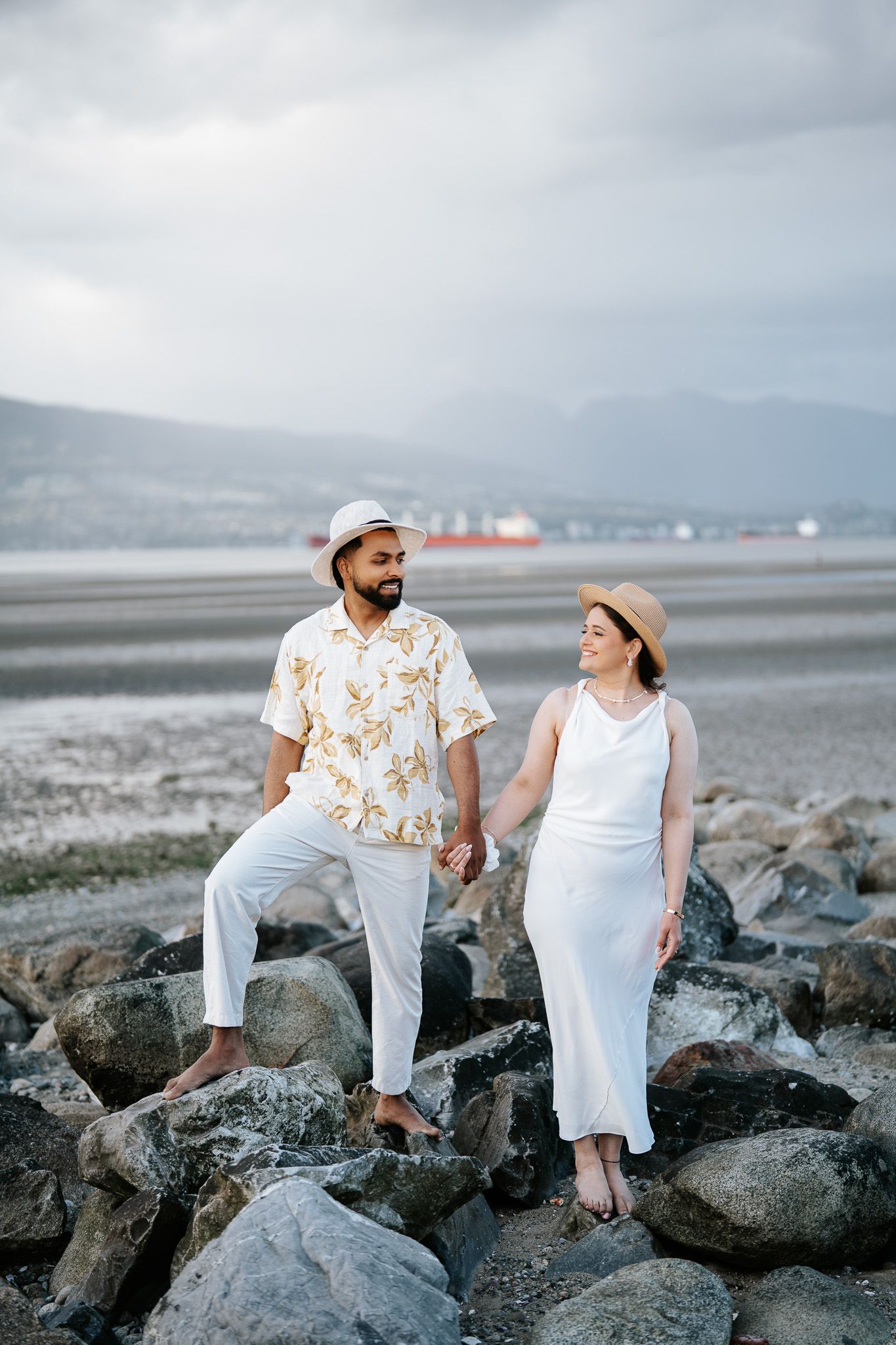 Couple in white outfits and straw hats holding hands while walking on rocky beach with mountains behind.