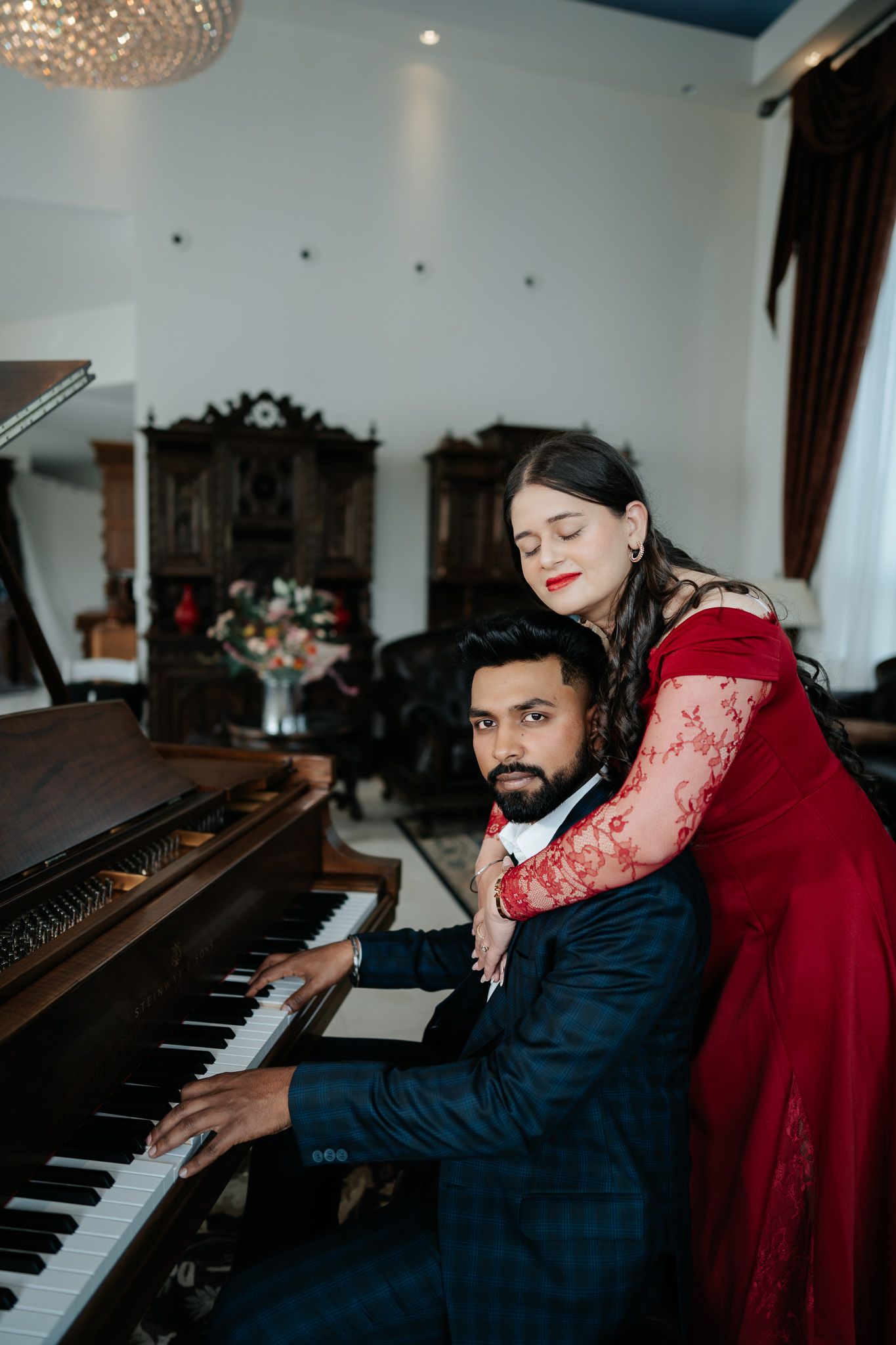 Couple in formal attire sharing romantic moment at a grand piano, woman in red lace gown embracing man.