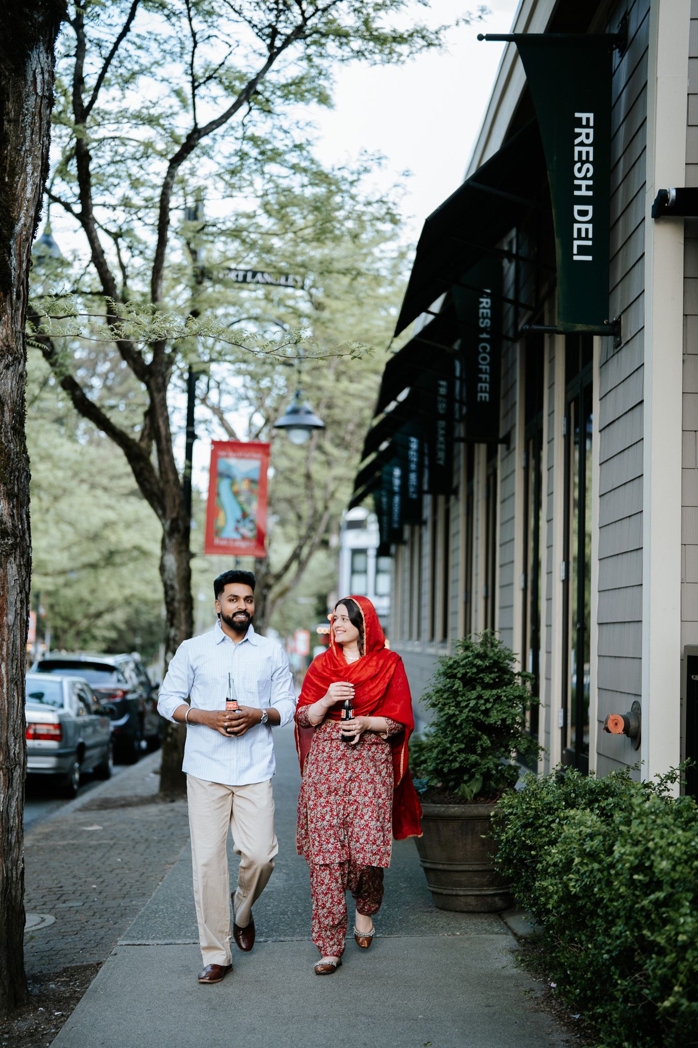Couple strolling on urban sidewalk past Fresh Deli shop, woman in red traditional Pakistani outfit.