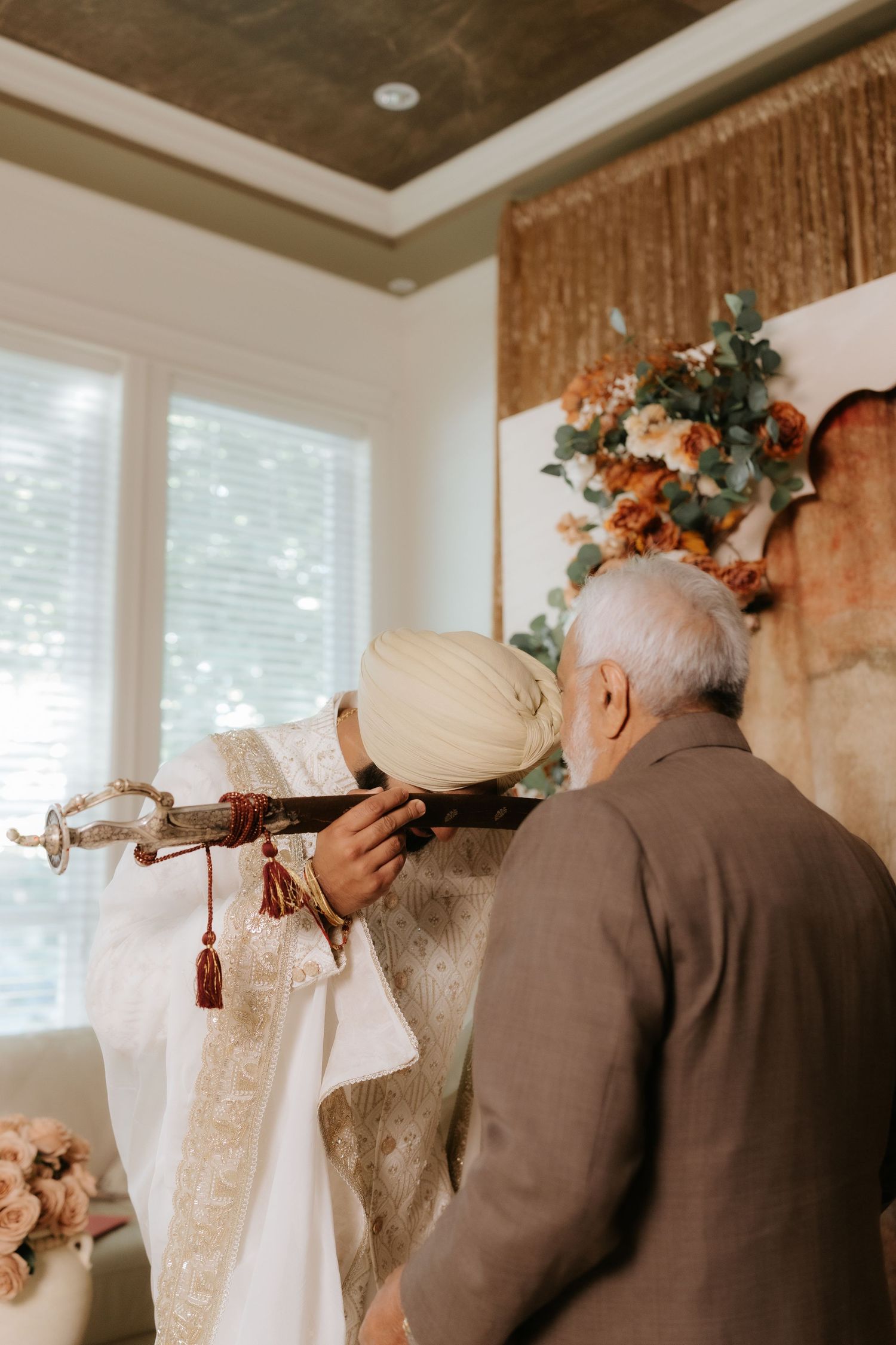 A Sikh groom in white sherwani and turban holds a ceremonial sword during a traditional wedding ritual indoors.