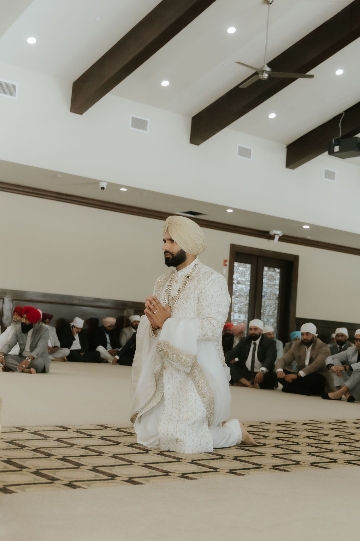 Sikh groom in white sherwani and red turban praying on knees at Gurdwara wedding ceremony.