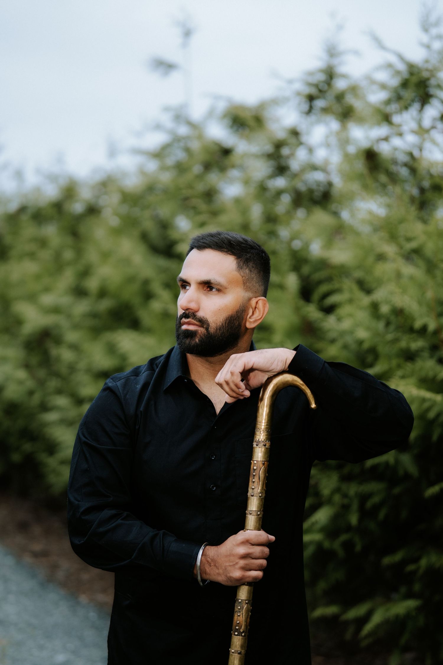 A bearded man in black shirt leans on ornate golden cane outdoors surrounded by lush green trees.