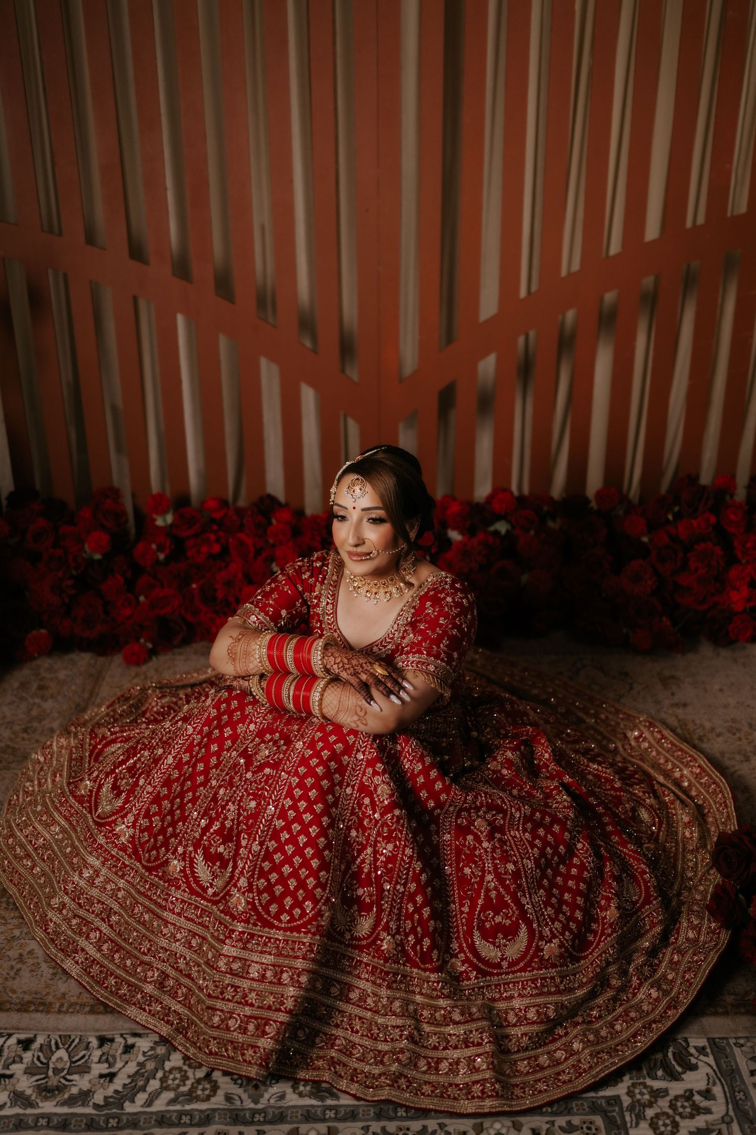 Indian bride in red embroidered lehenga with gold jewelry sits gracefully, surrounded by red roses in elegant bridal portrait.