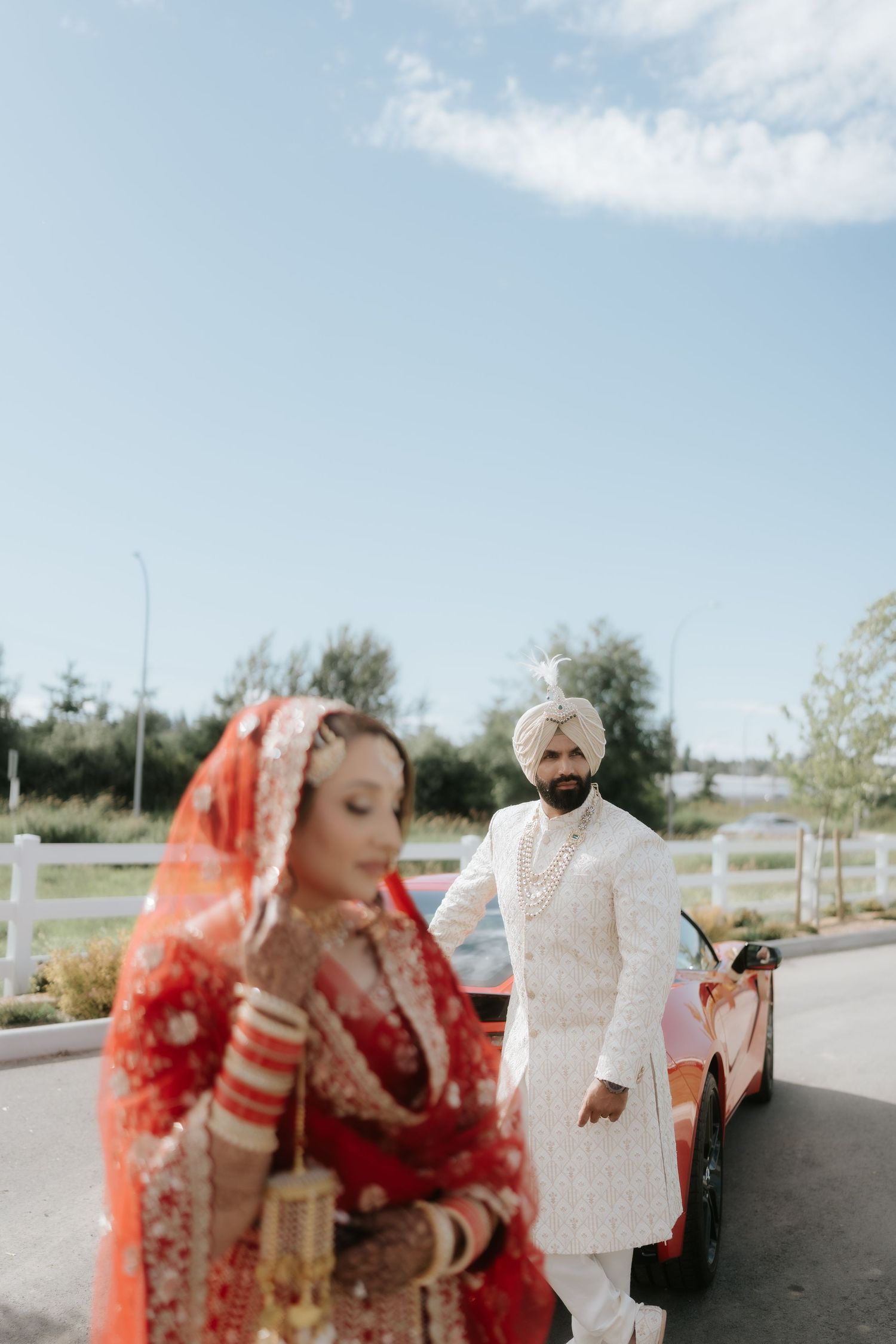 Sikh bride in red lehenga and groom in white sherwani posing outdoors near a red car on their wedding day.