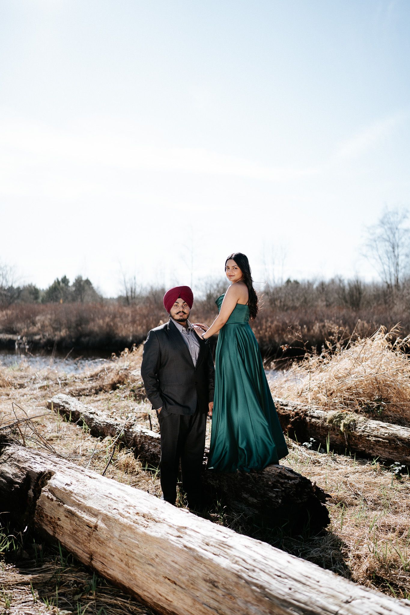 A couple in formal attire sits together on a snowy log in a winter landscape during a photo session.