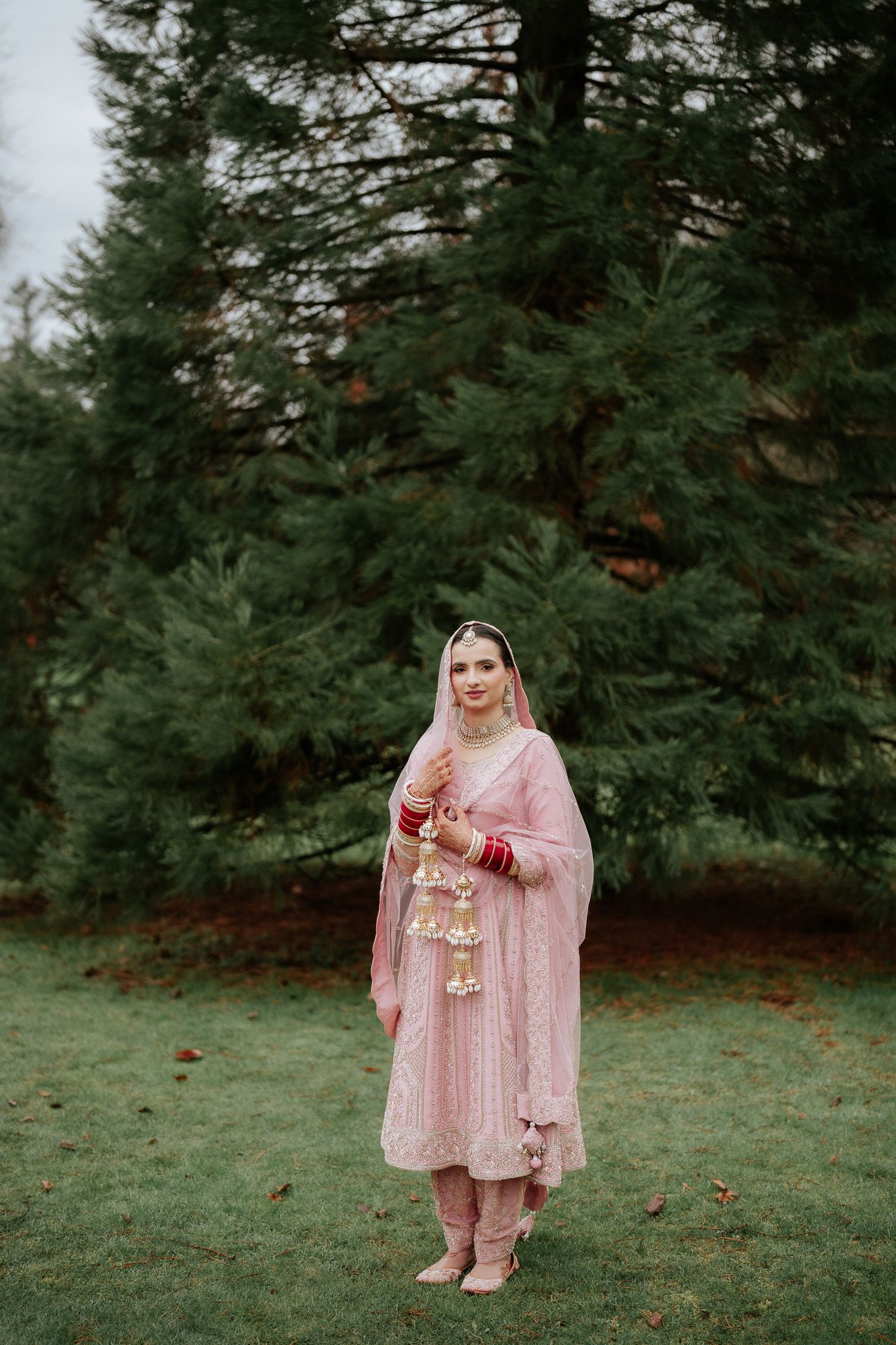 A bride in a pink traditional dress and veil poses outdoors against a backdrop of evergreen trees.