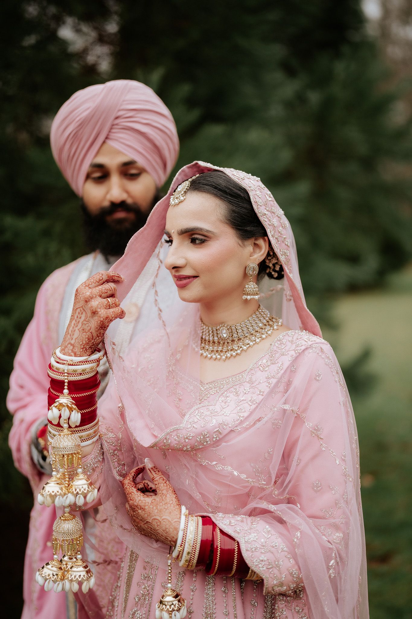 Traditional Sikh wedding couple in pink outfits with bride wearing ornate jewelry and henna during outdoor ceremony.
