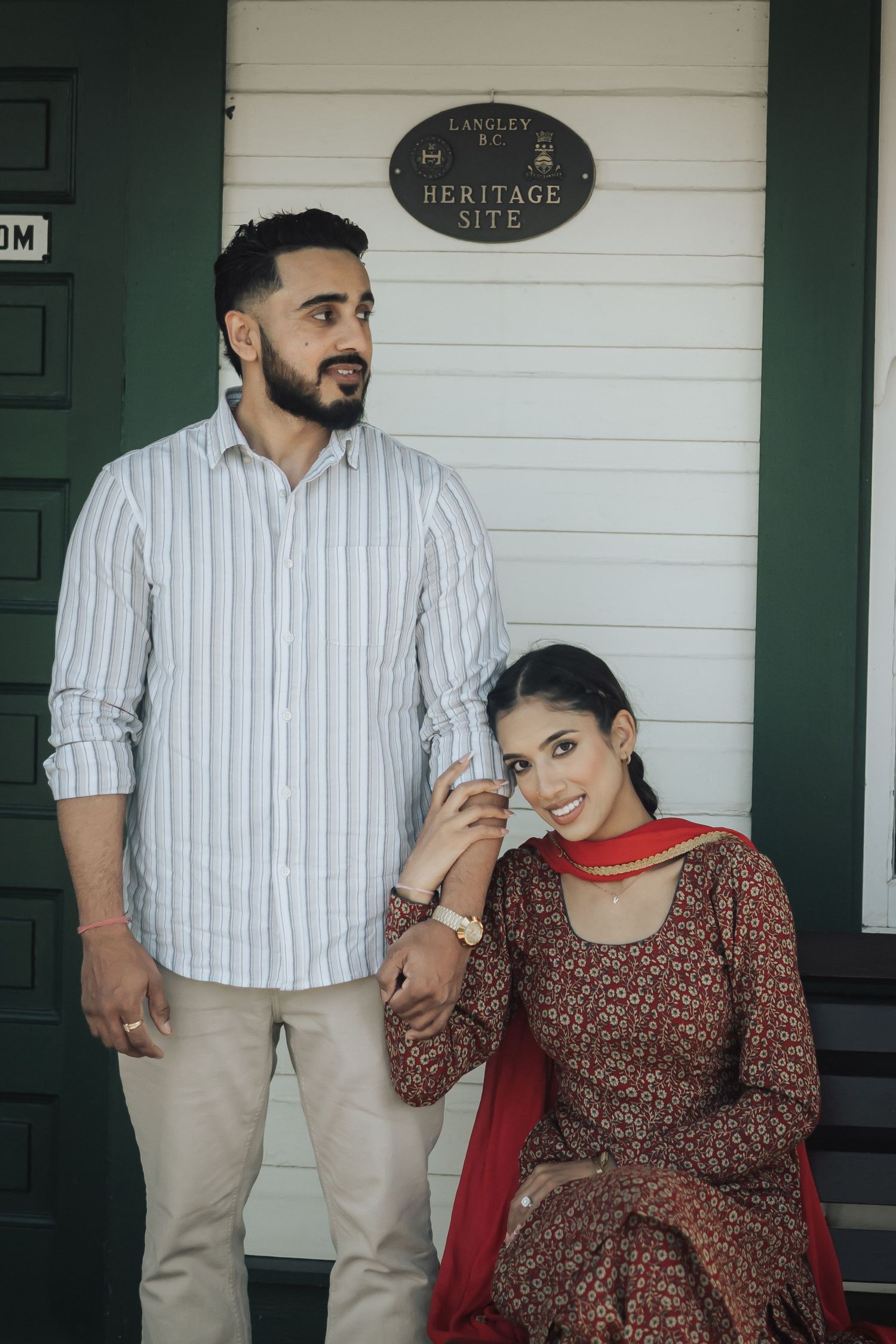 A couple poses outside a heritage building, with the person standing wearing a striped shirt and the other in a maroon dress.