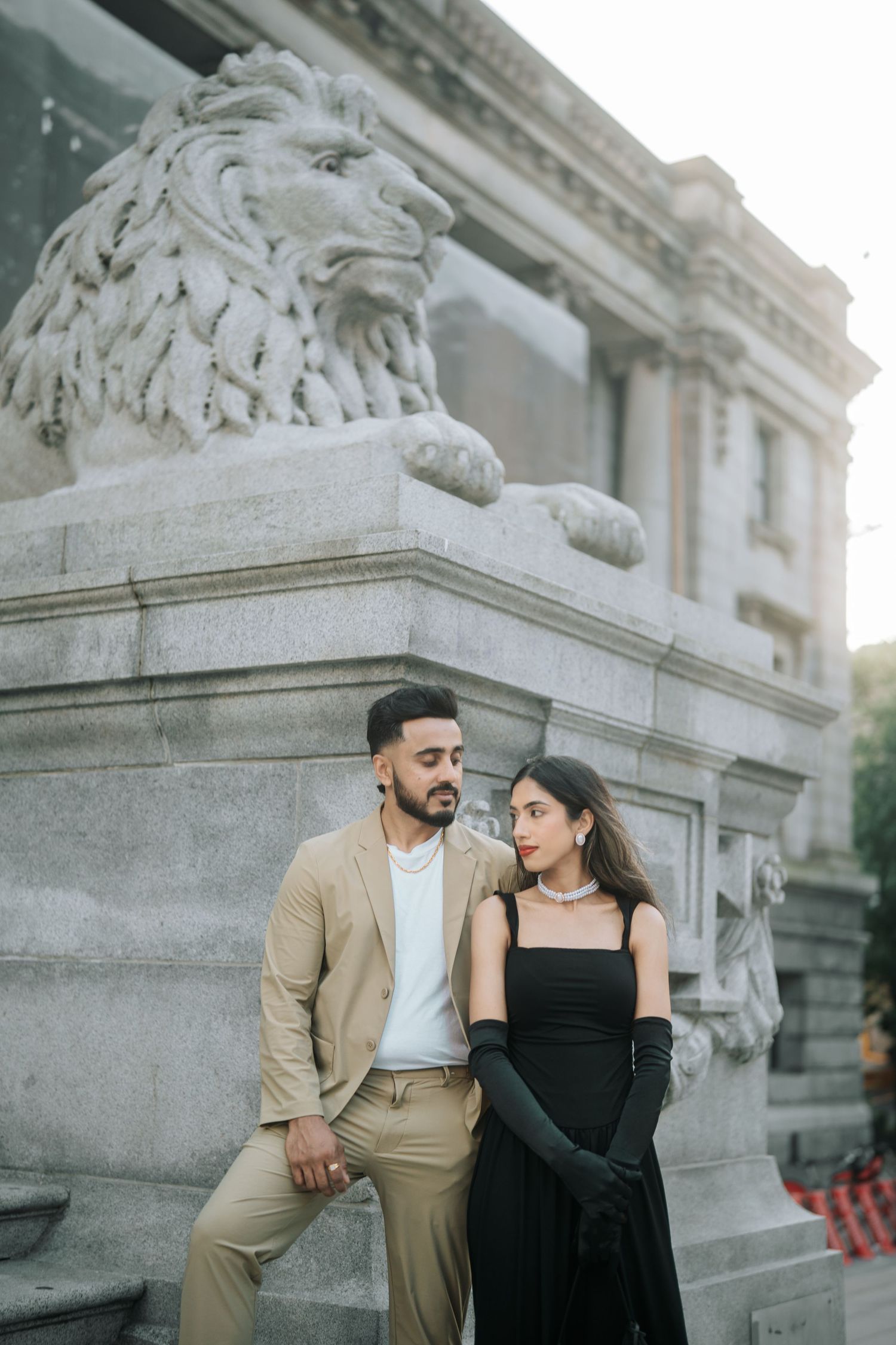 A couple in formal attire poses together near an ornate stone monument with a carved lion statue in an elegant setting.
