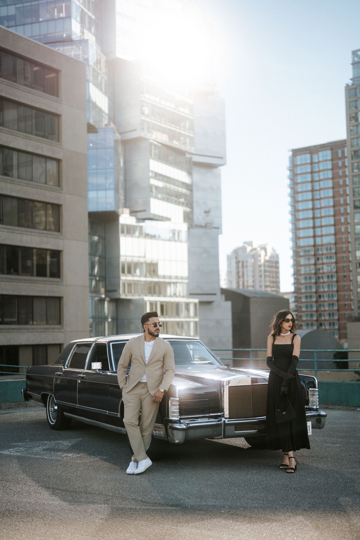A stylish couple poses with a vintage black limousine on a modern urban rooftop with city buildings in the background.