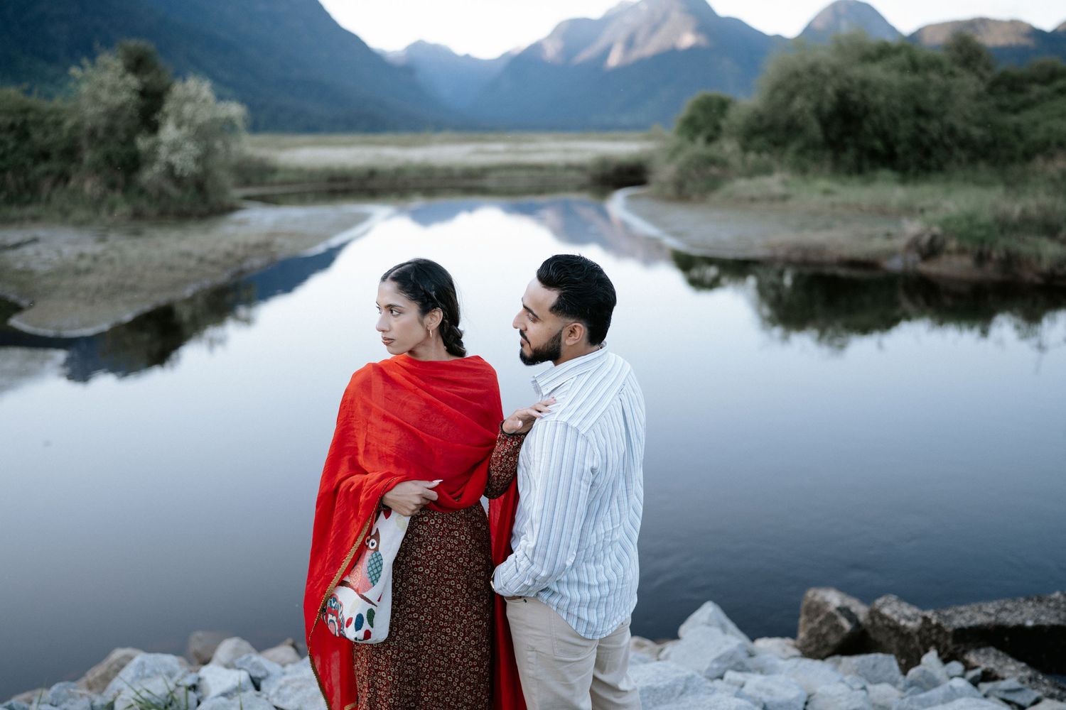 A couple in red and white outfits sit together by a serene mountain lake surrounded by majestic peaks.