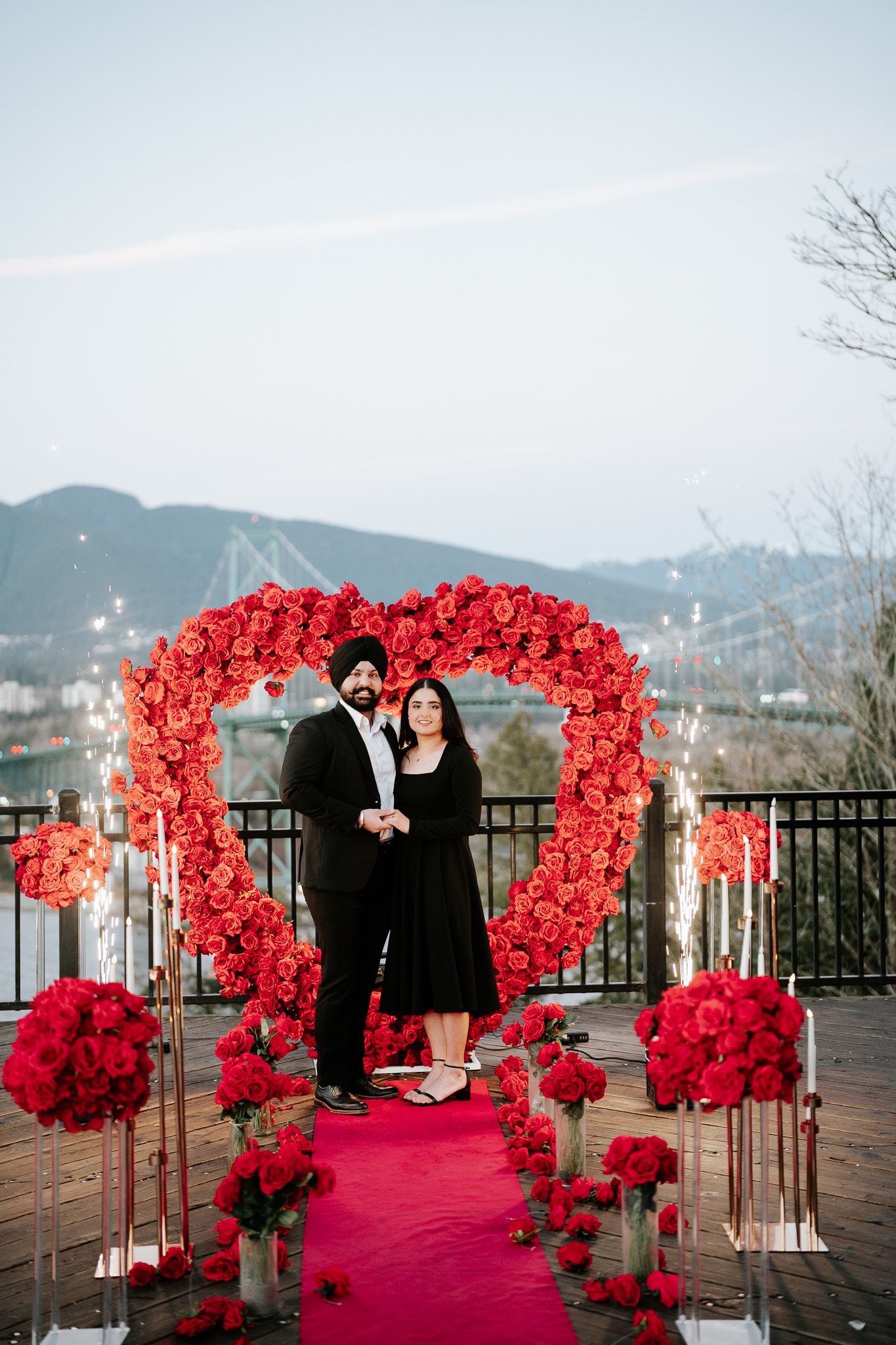 A couple stands beneath a large heart-shaped red floral arch on a red carpet with mountains in the background.
