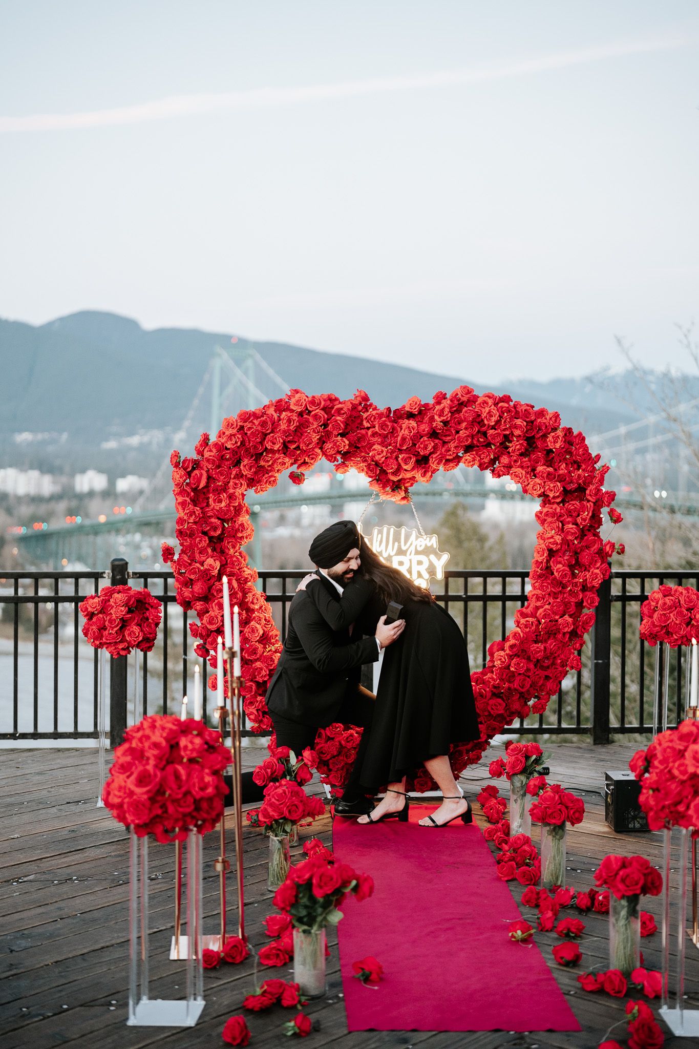 A romantic proposal scene unfolds under a red rose arch on a deck overlooking mountains at sunset.