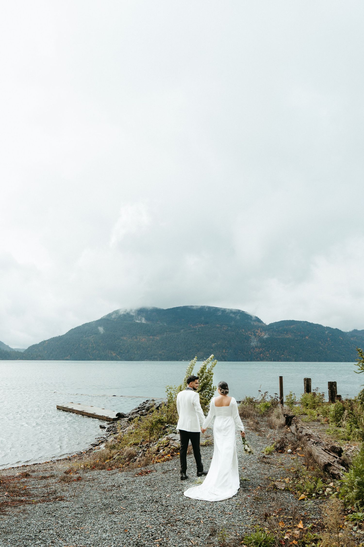 Couple walking along rocky beach shore with misty mountains and cloudy skies in Alaska.