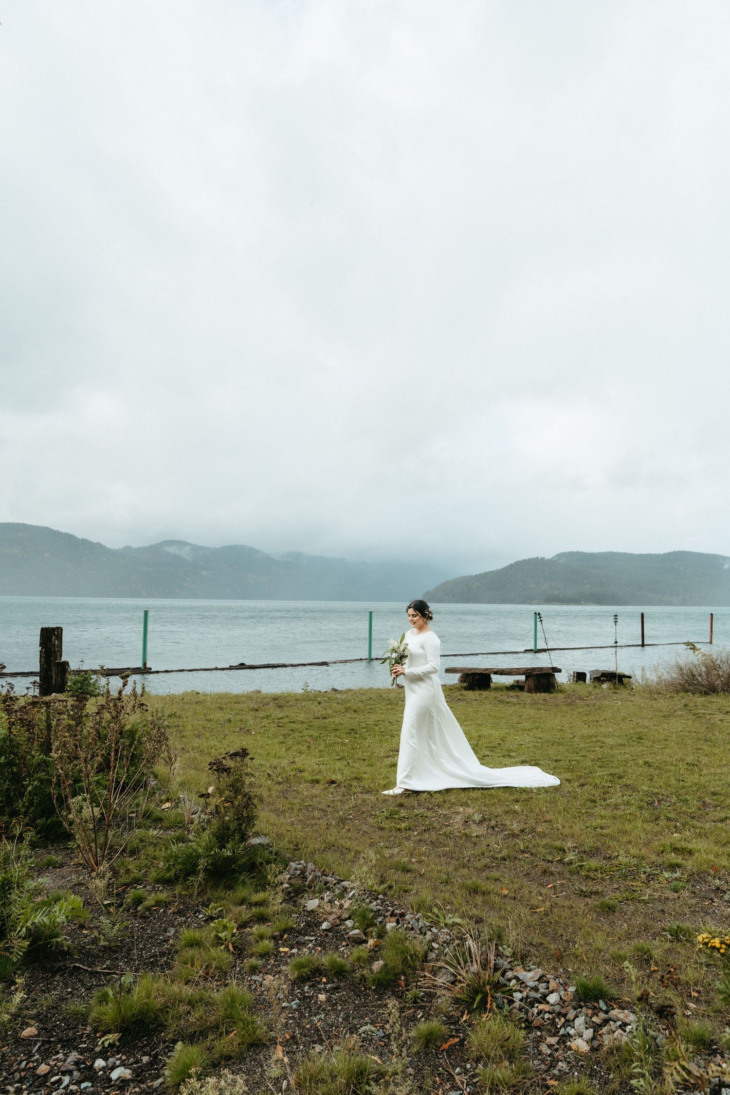 A figure in a flowing white wedding dress stands on a grassy cliff overlooking a misty bay with mountains in the distance.