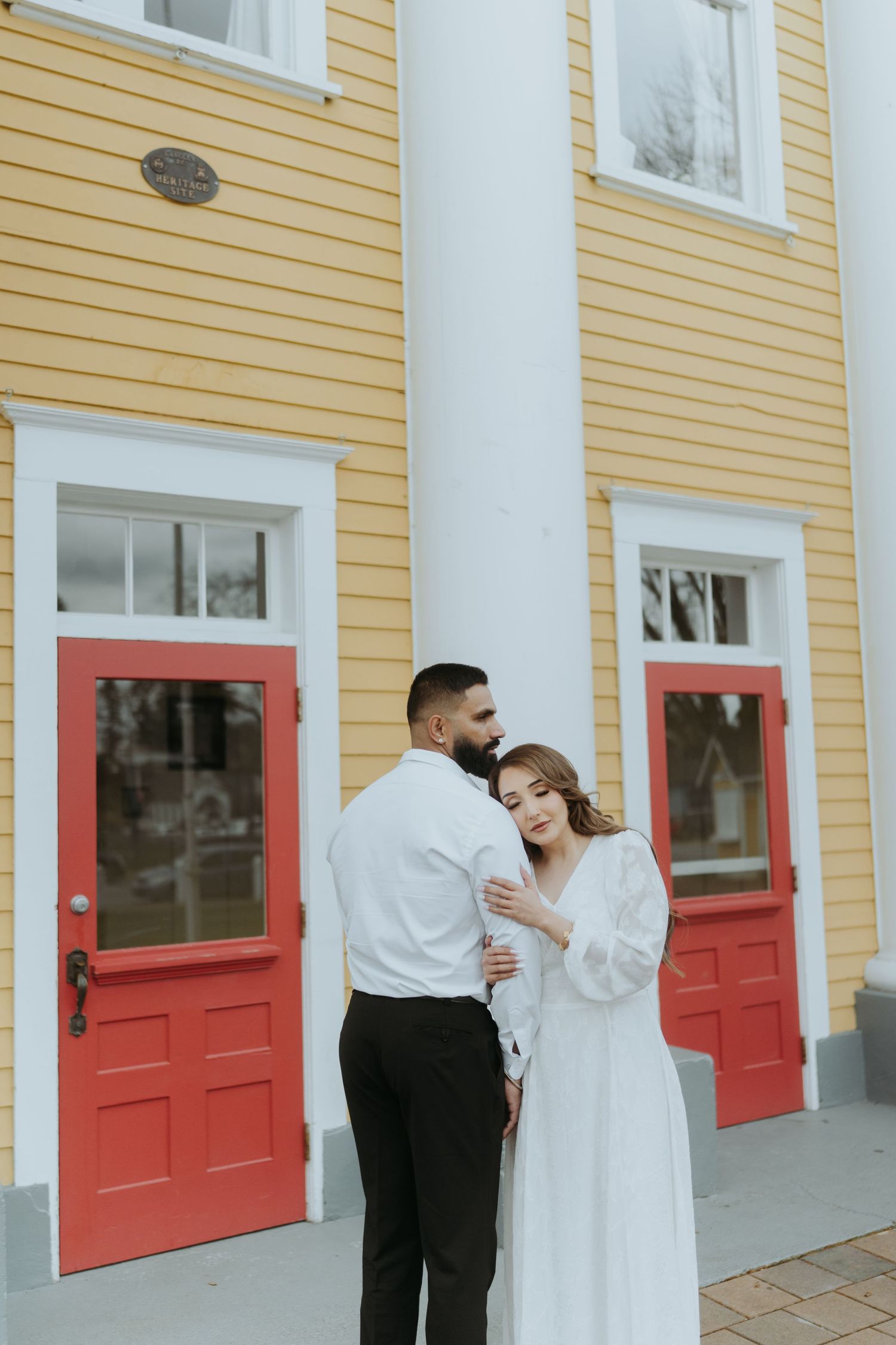 A couple embraces in front of a yellow building with bright red doors and white trim.