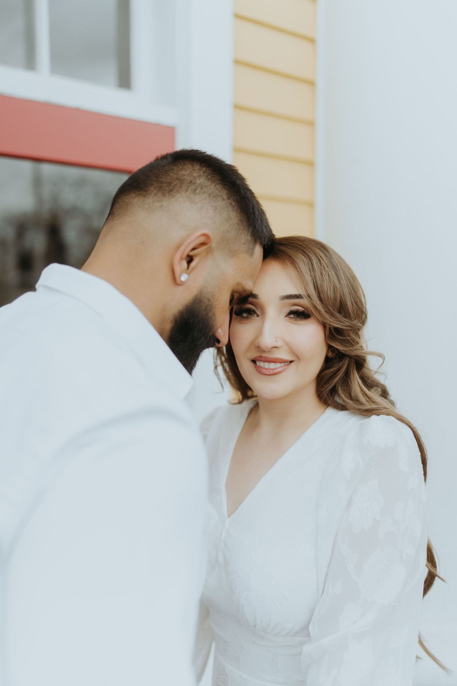 A couple dressed in white embraces while smiling against a building exterior.