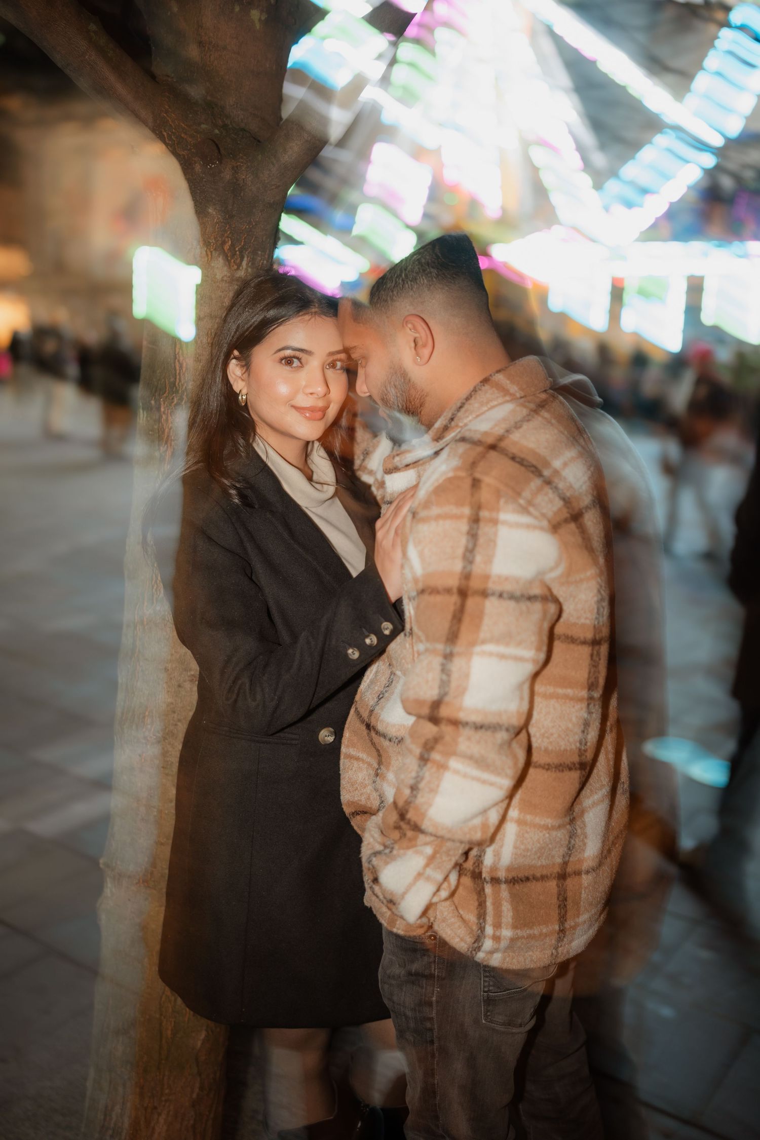 A romantic couple embraces under twinkling lights at dusk, with the woman in a black coat and man in plaid.