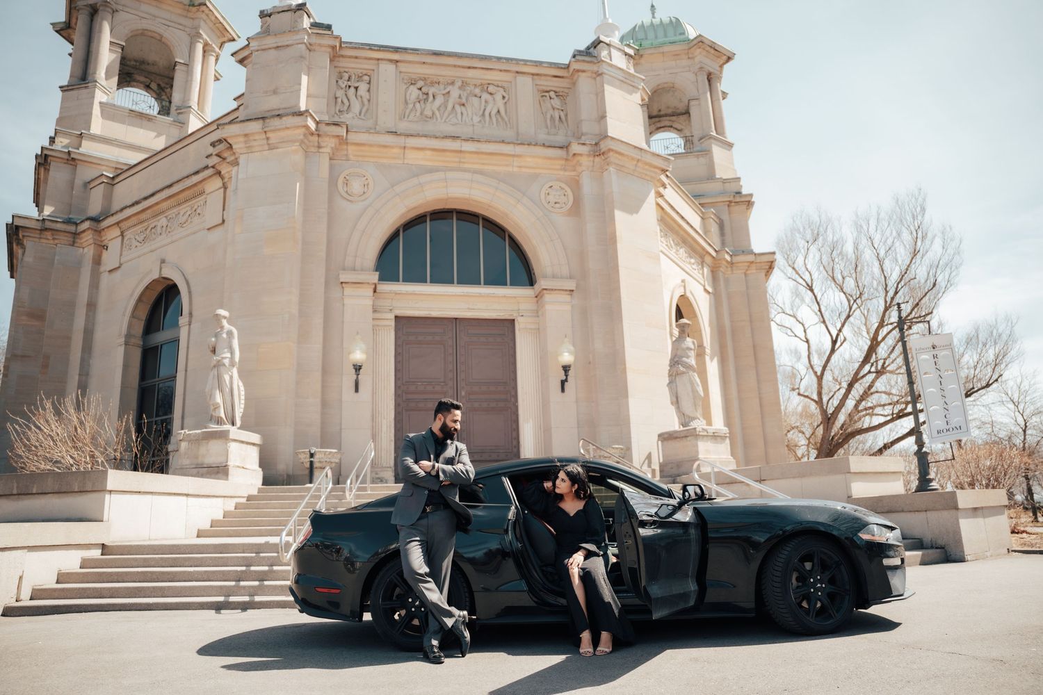 A group poses by a luxury black car in front of an elegant beige classical building with arched entrance and towers.