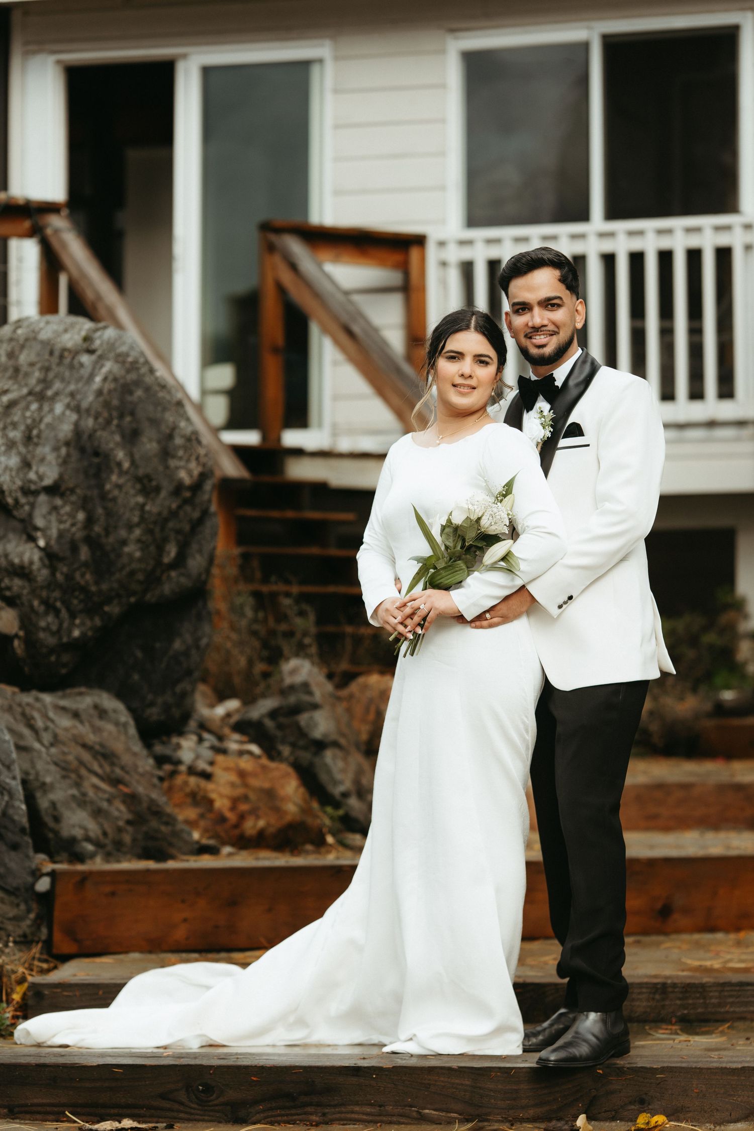 A couple in wedding attire sits together on rustic wooden steps outside a white house.
