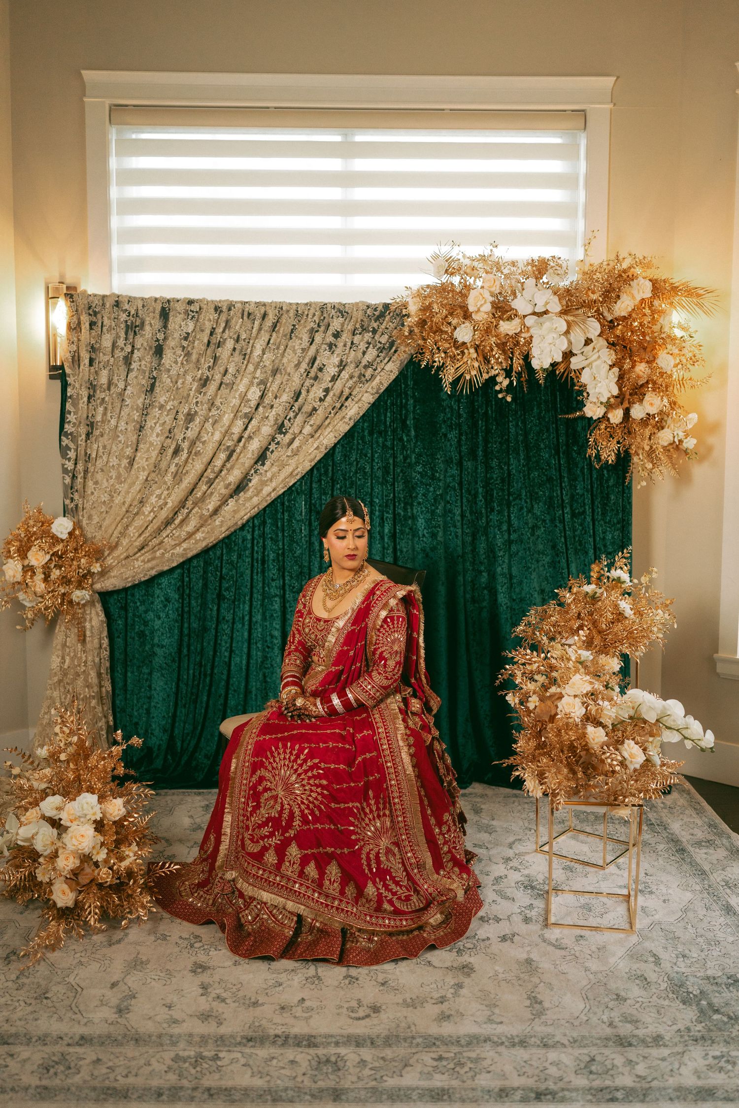 Indian bride in red lehenga sits before green velvet backdrop with gold and white floral arrangements.
