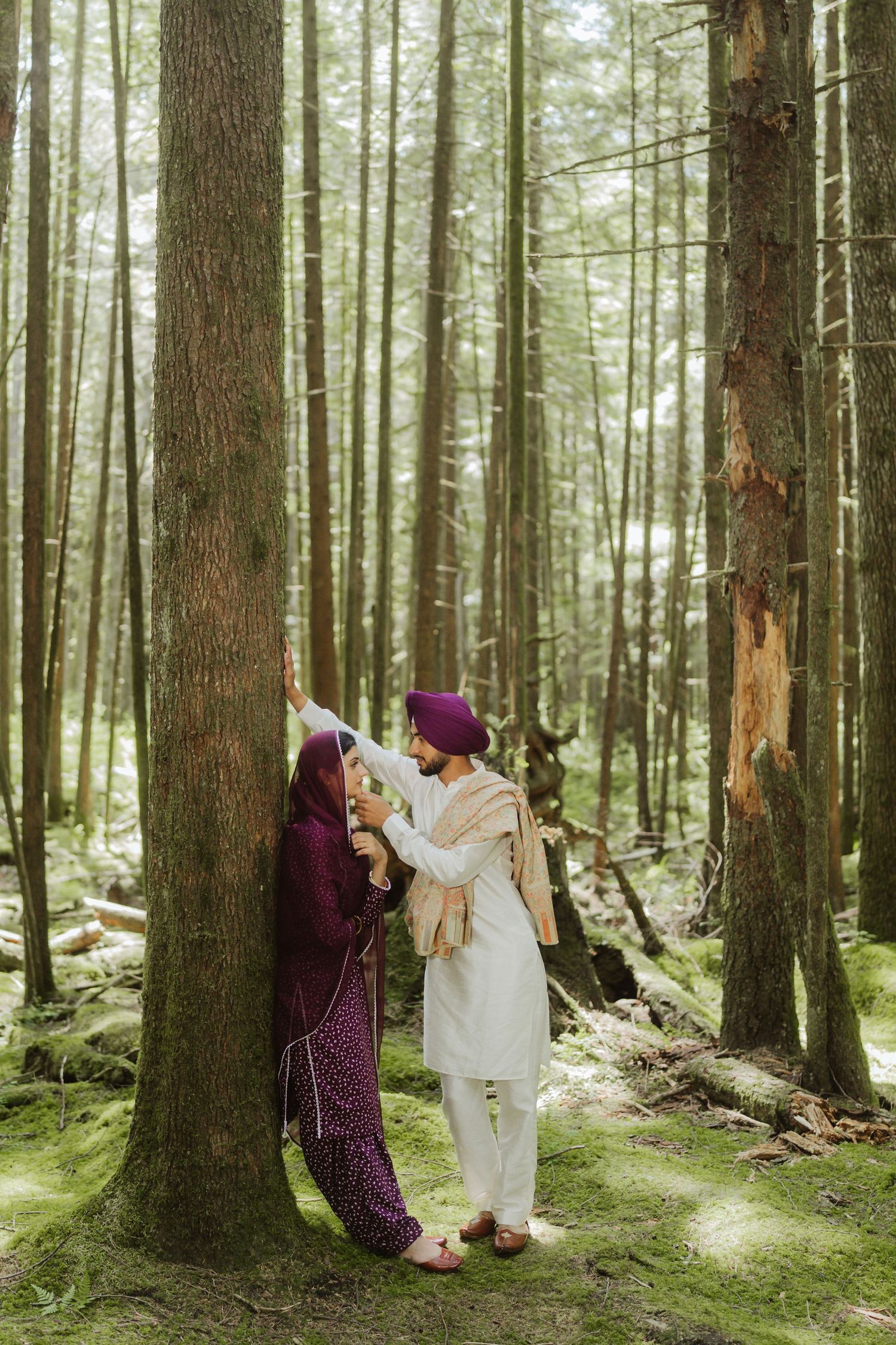 Sikh couple standing by tall forest tree, woman in maroon suit facing man in white kurta and turban.