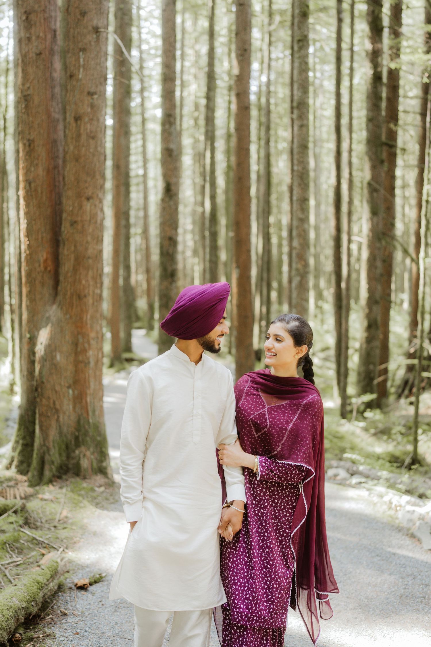 Sikh couple walking hand in hand through a forest, wearing traditional white kurta and burgundy salwar suit.