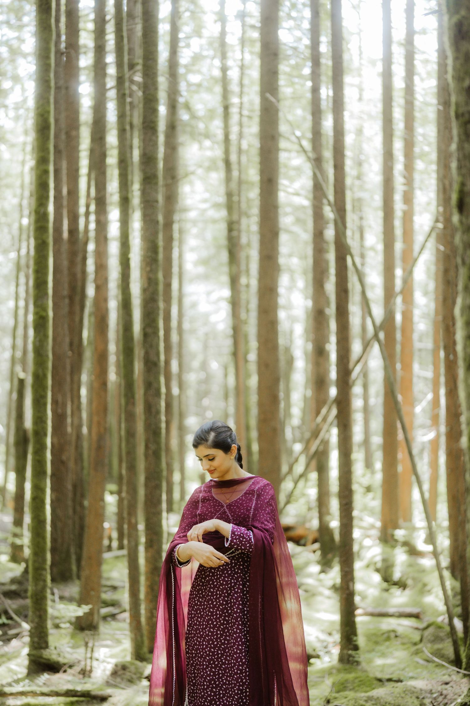 Woman in burgundy salwar kameez with dupatta stands gracefully among tall forest trees in soft natural light.