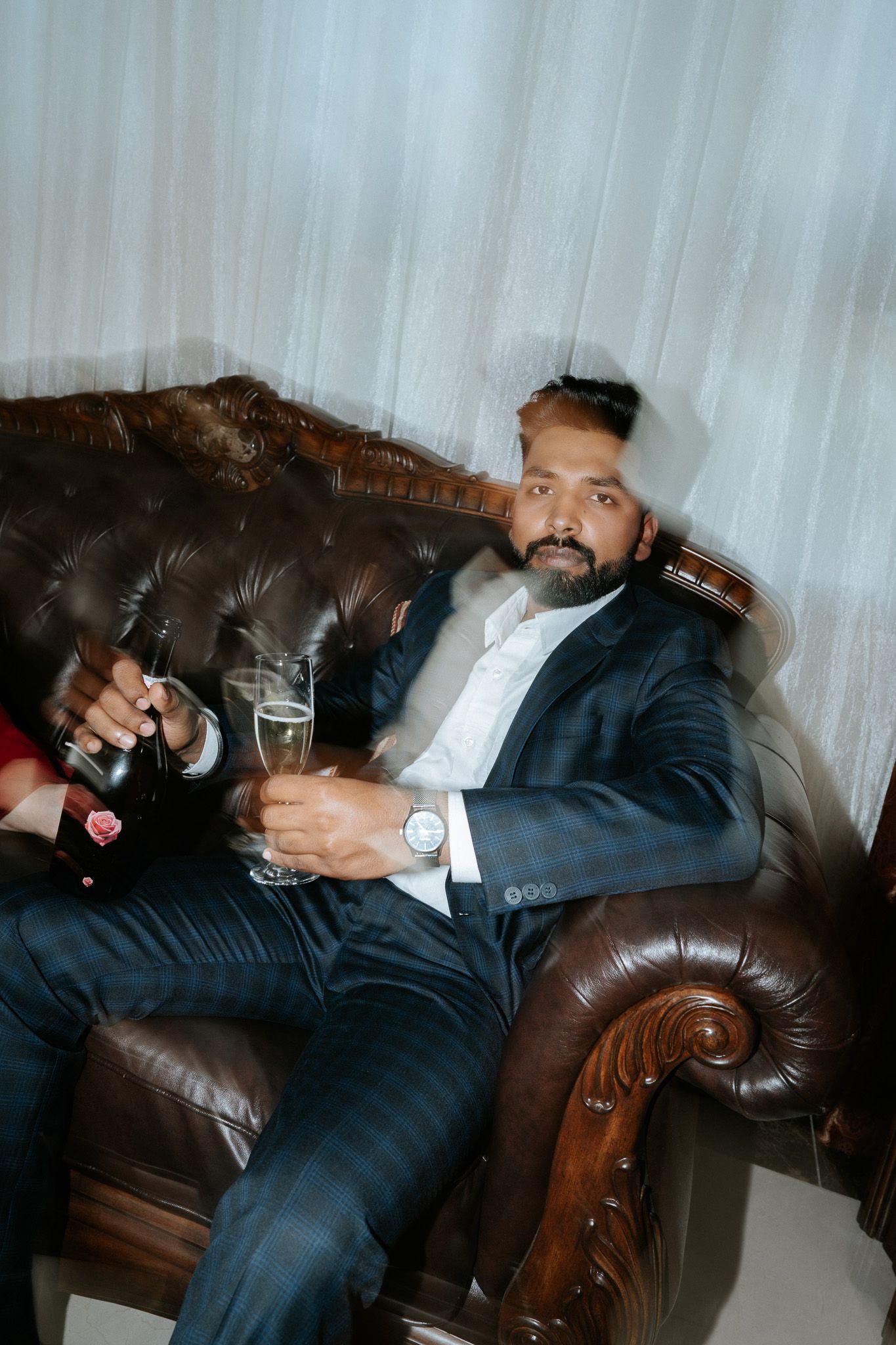 Man in navy suit relaxing on leather sofa holding champagne glass at formal event.